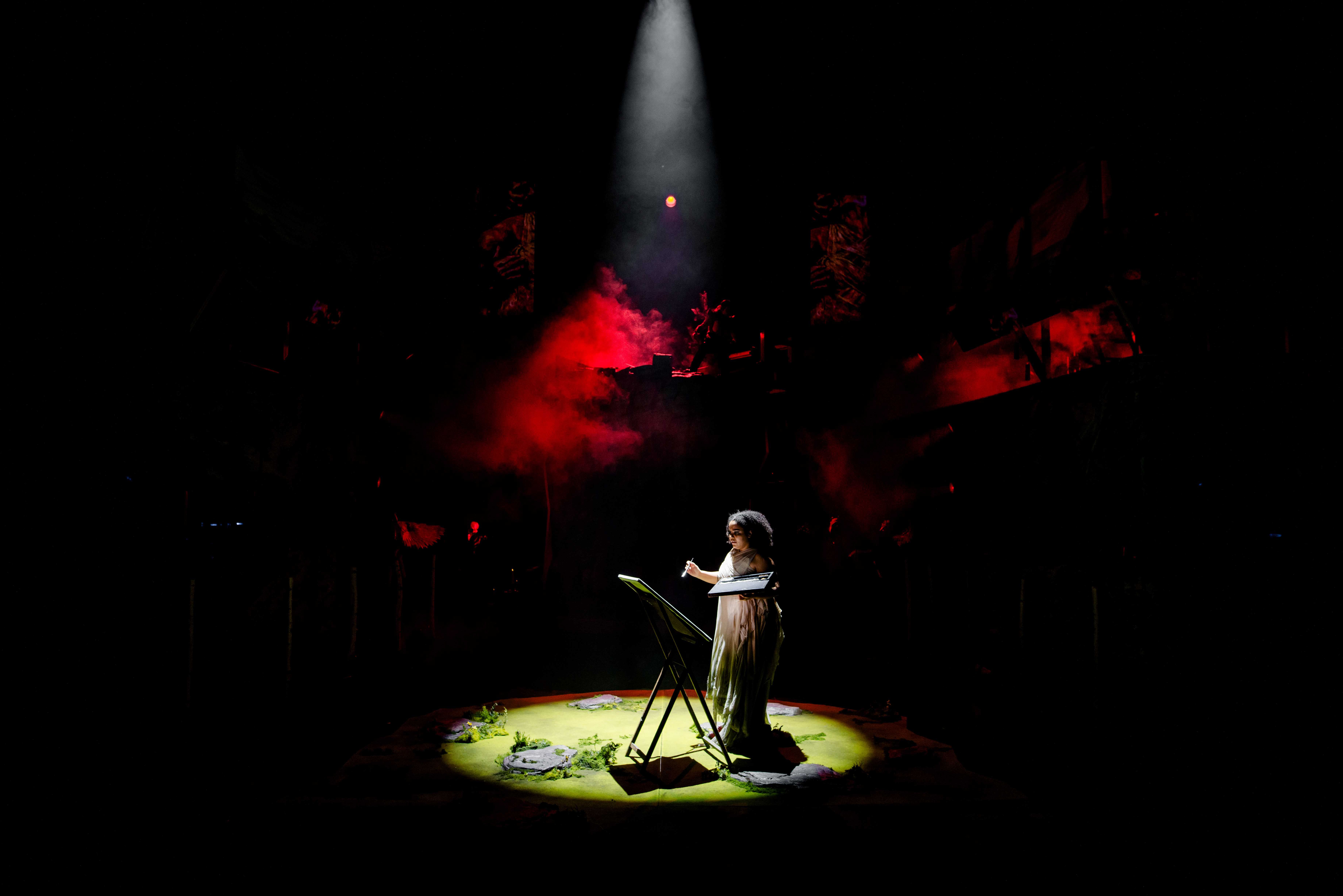 Kvinne i lys kjole dirigerer foran et staffeli og maler et bilde i en scenesirkel med røyk og rød belysning rundt seg.