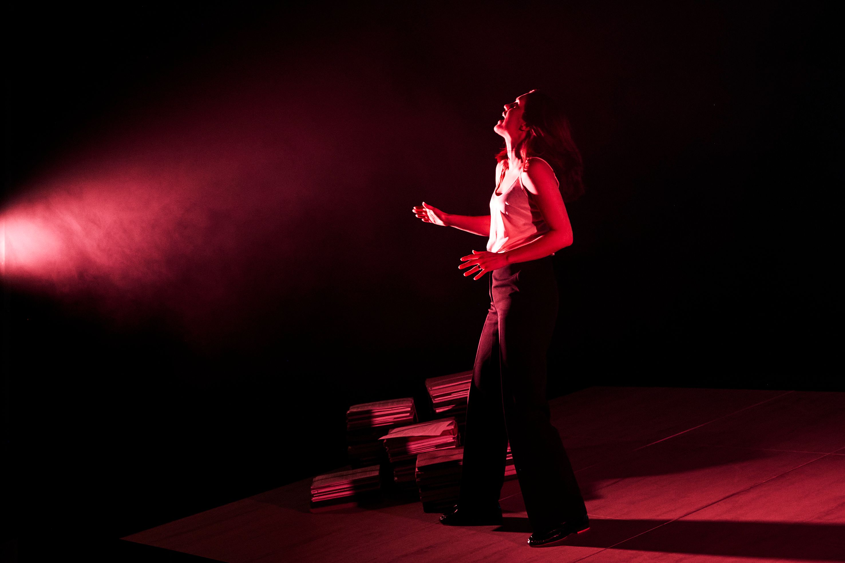 En kvinne i hvit topp og svarte bukser står i rødt lys med hodet bakover og armene løftet.