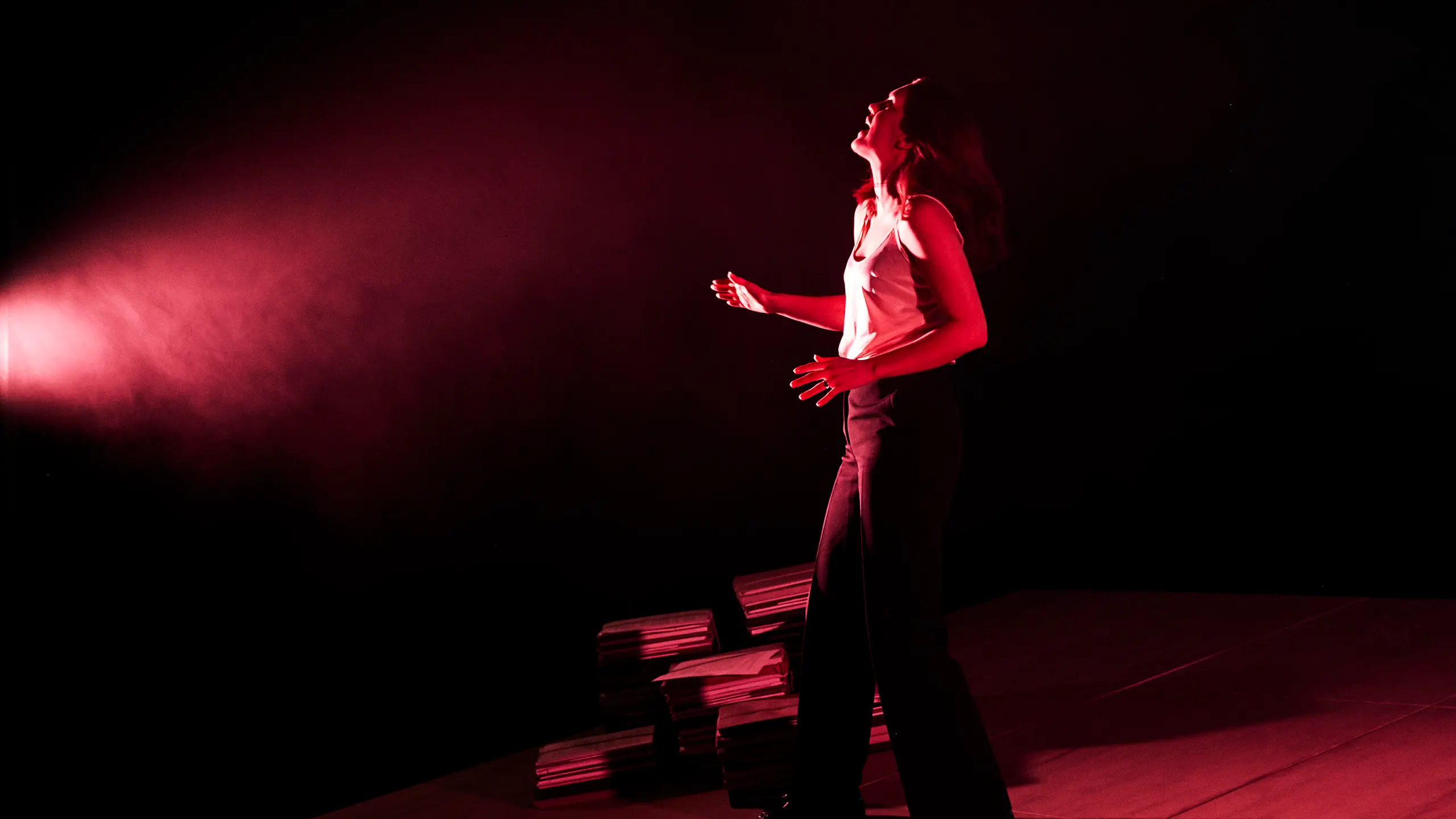 En kvinne i hvit topp og svarte bukser står i rødt lys med hodet bakover og armene løftet.