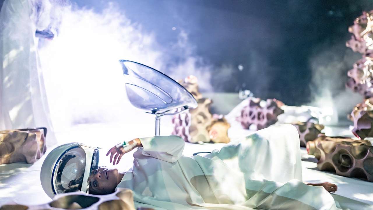 Person i hvit drakt ligger på gulvet med romhjelm, omgitt av røyk, futuristisk stol og organiske skulpturer i bakgrunnen