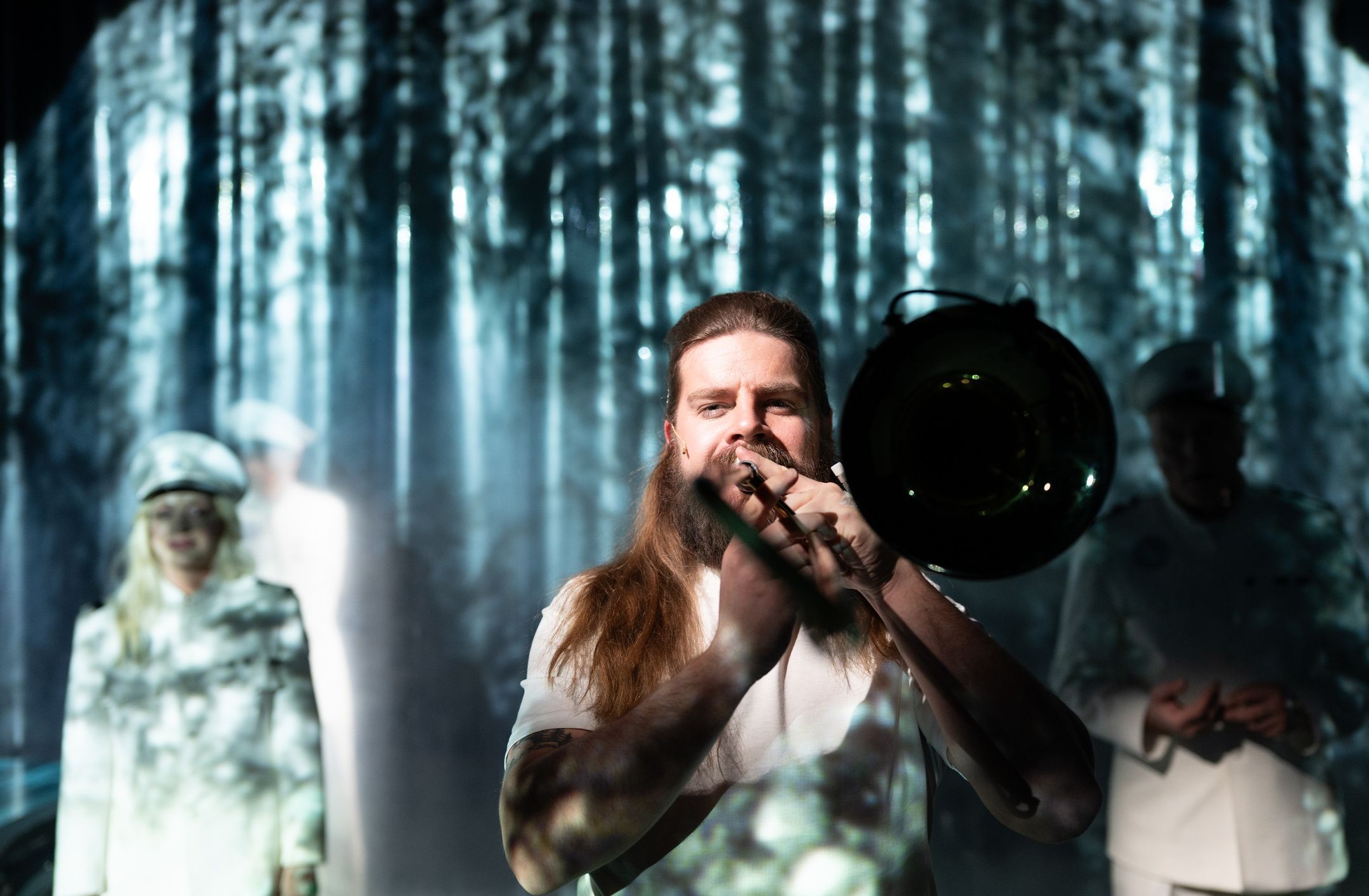 Langhåret person med skjegg spiller trombone i forgrunnen, uniformerte personer står uklart bak