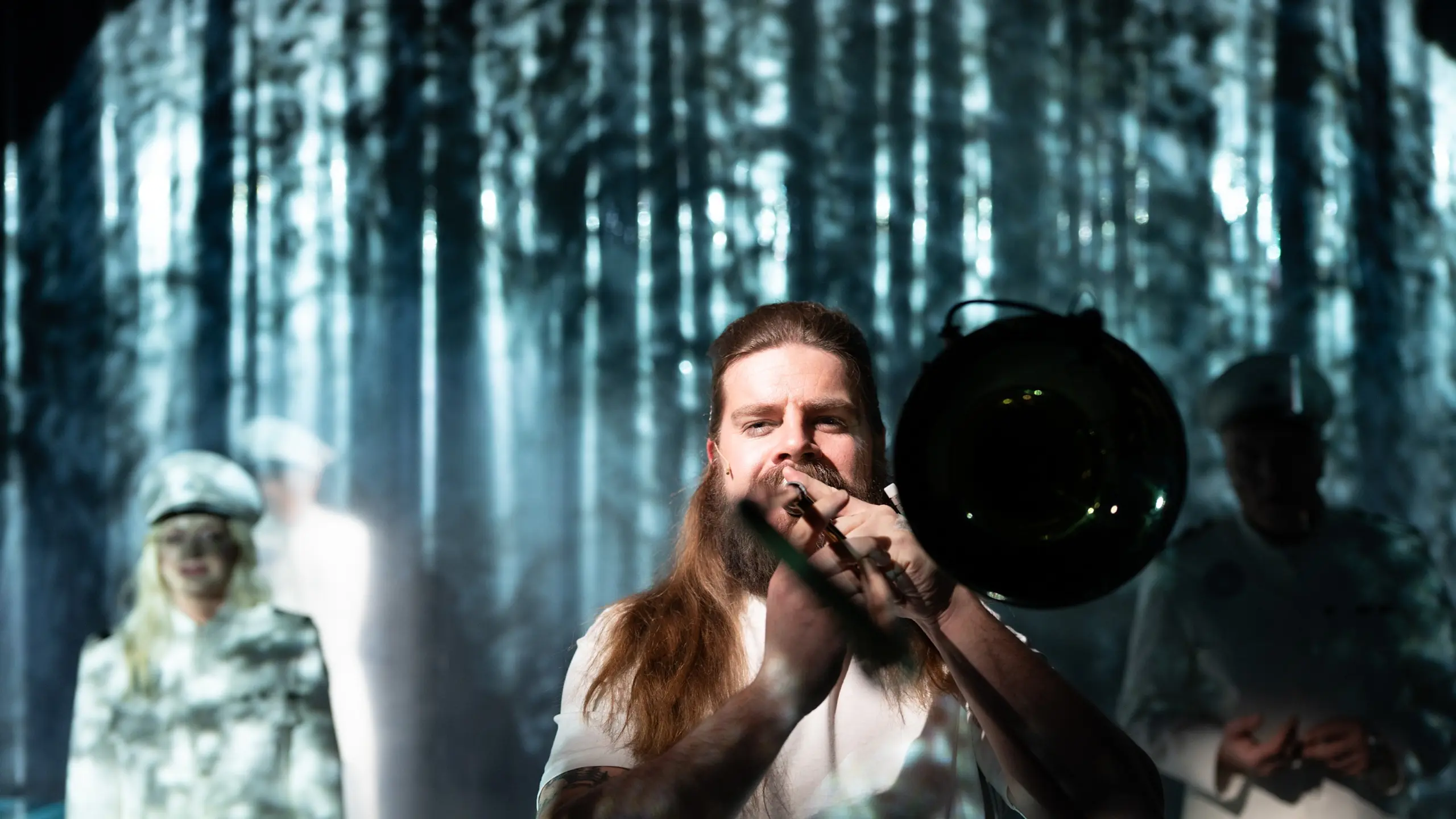 Langhåret person med skjegg spiller trombone i forgrunnen, uniformerte personer står uklart bak