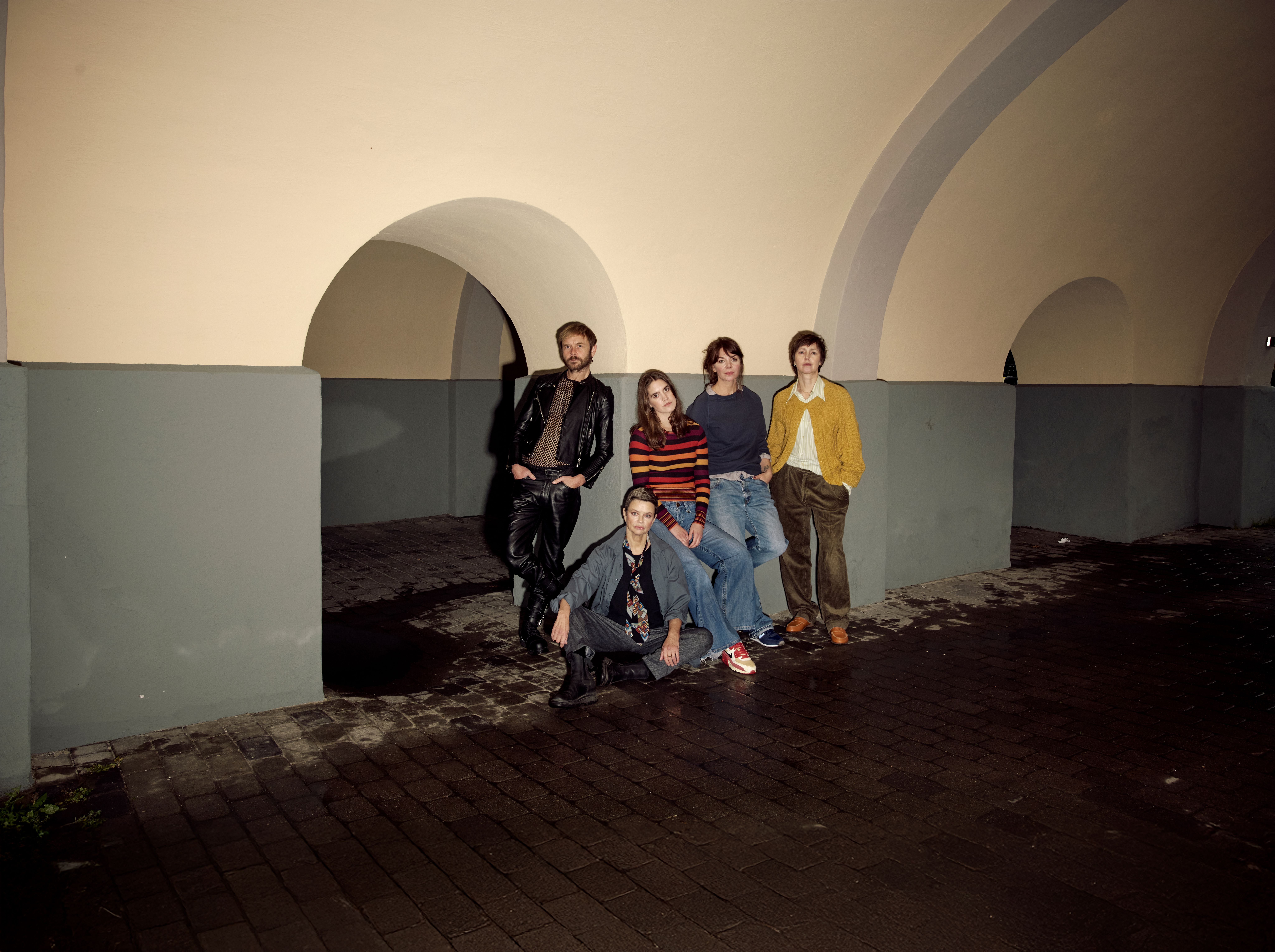 Gruppe med fire kvinner og en mann i en buegang. De er alvorlige, den ene kvinnen sitter, resten står og lener seg mot veggen