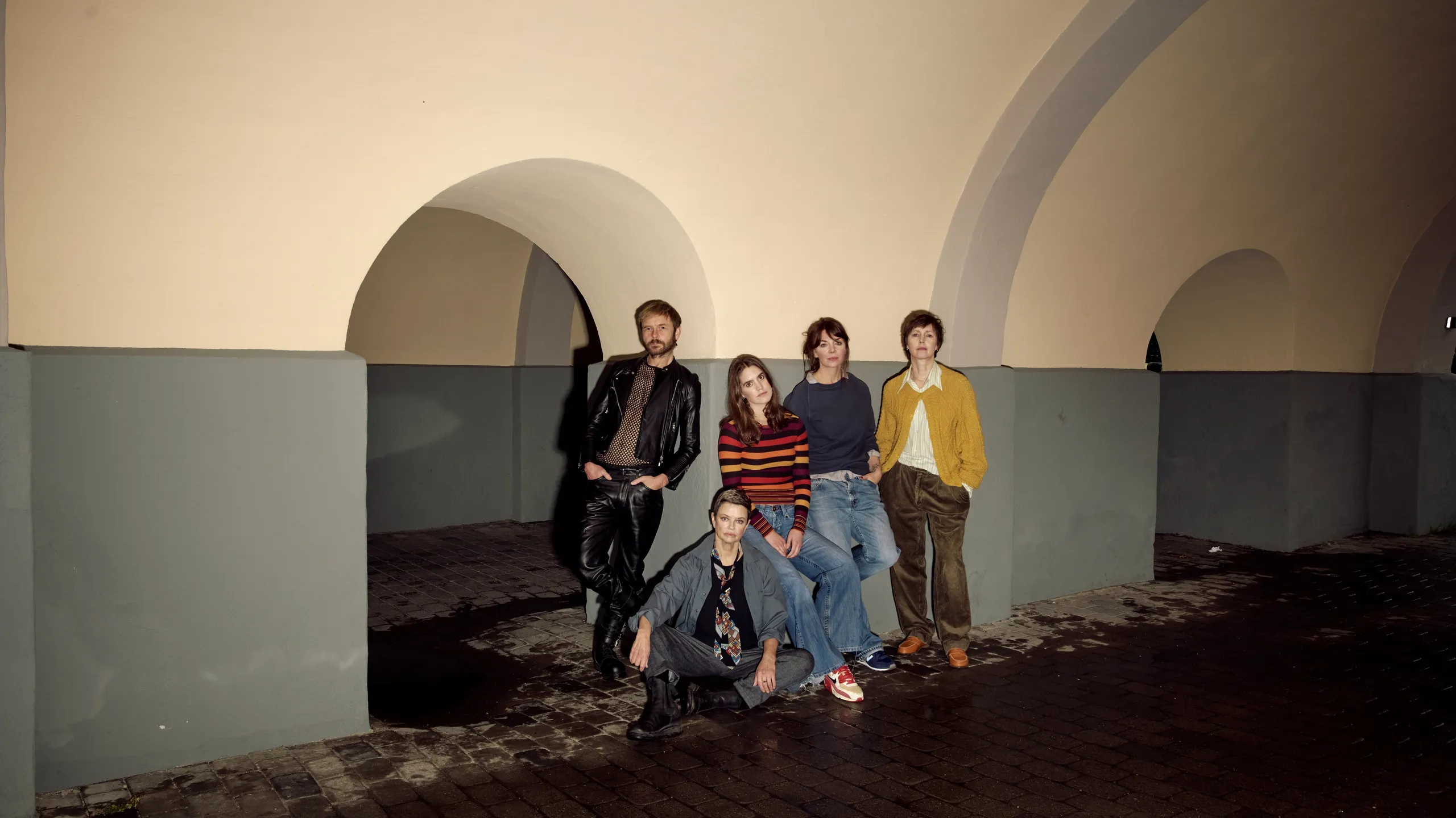 Gruppe med fire kvinner og en mann i en buegang. De er alvorlige, den ene kvinnen sitter, resten står og lener seg mot veggen