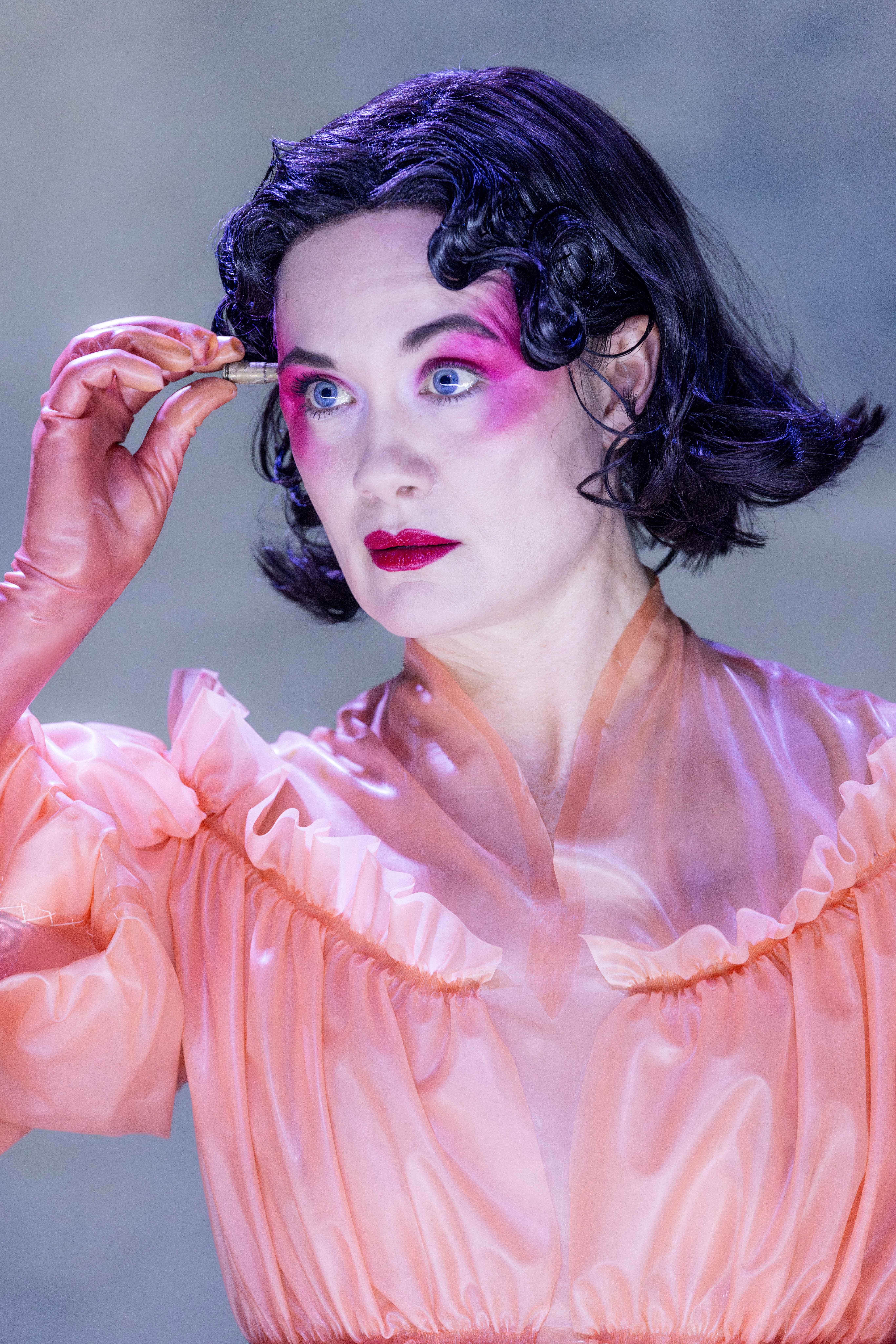 Person med kort mørkt hår, kraftig rosa øyenskygge og ferskenfarget kjole holder en liten patron foran pannen