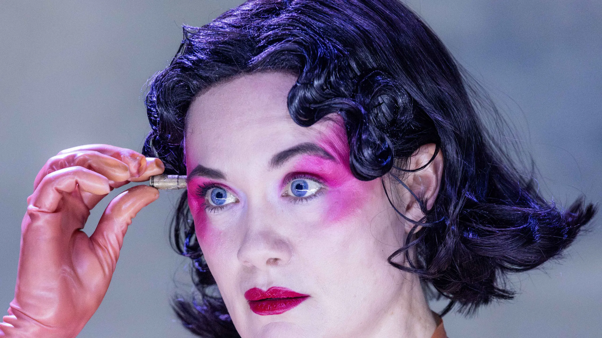 Person med kort mørkt hår, kraftig rosa øyenskygge og ferskenfarget kjole holder en liten patron foran pannen