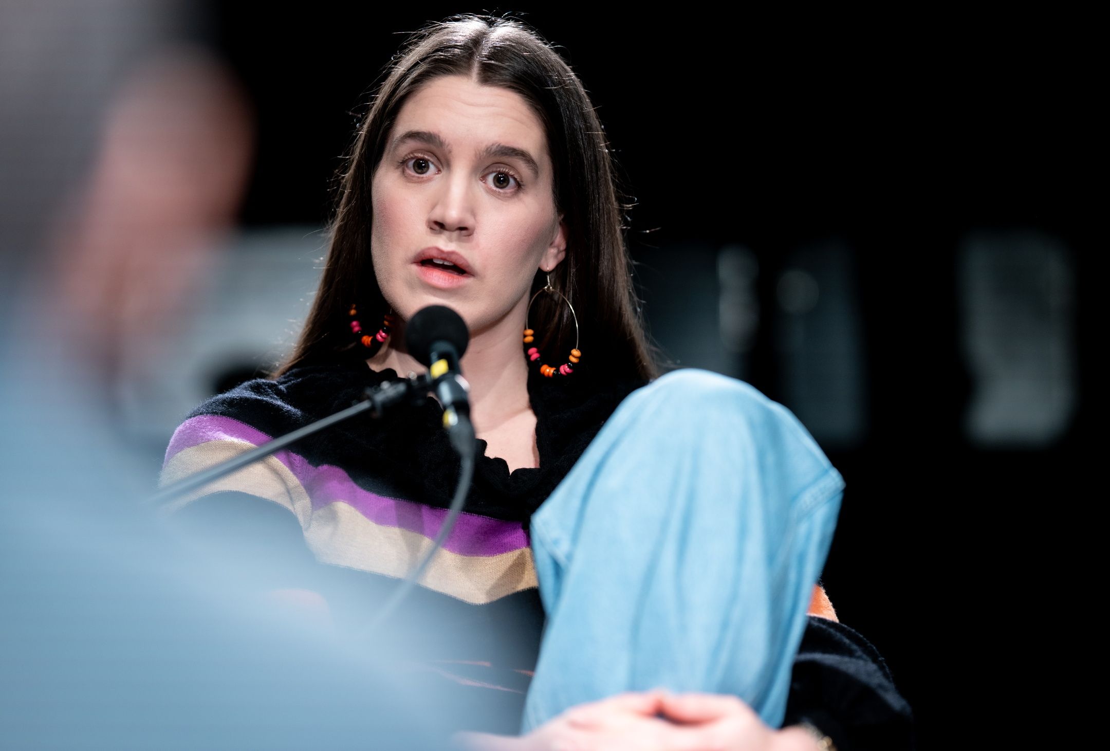 Person med langt mørkt hår, store runde øreringer og stripet genser snakker inn i mikrofon med oppmerksomt uttrykk.