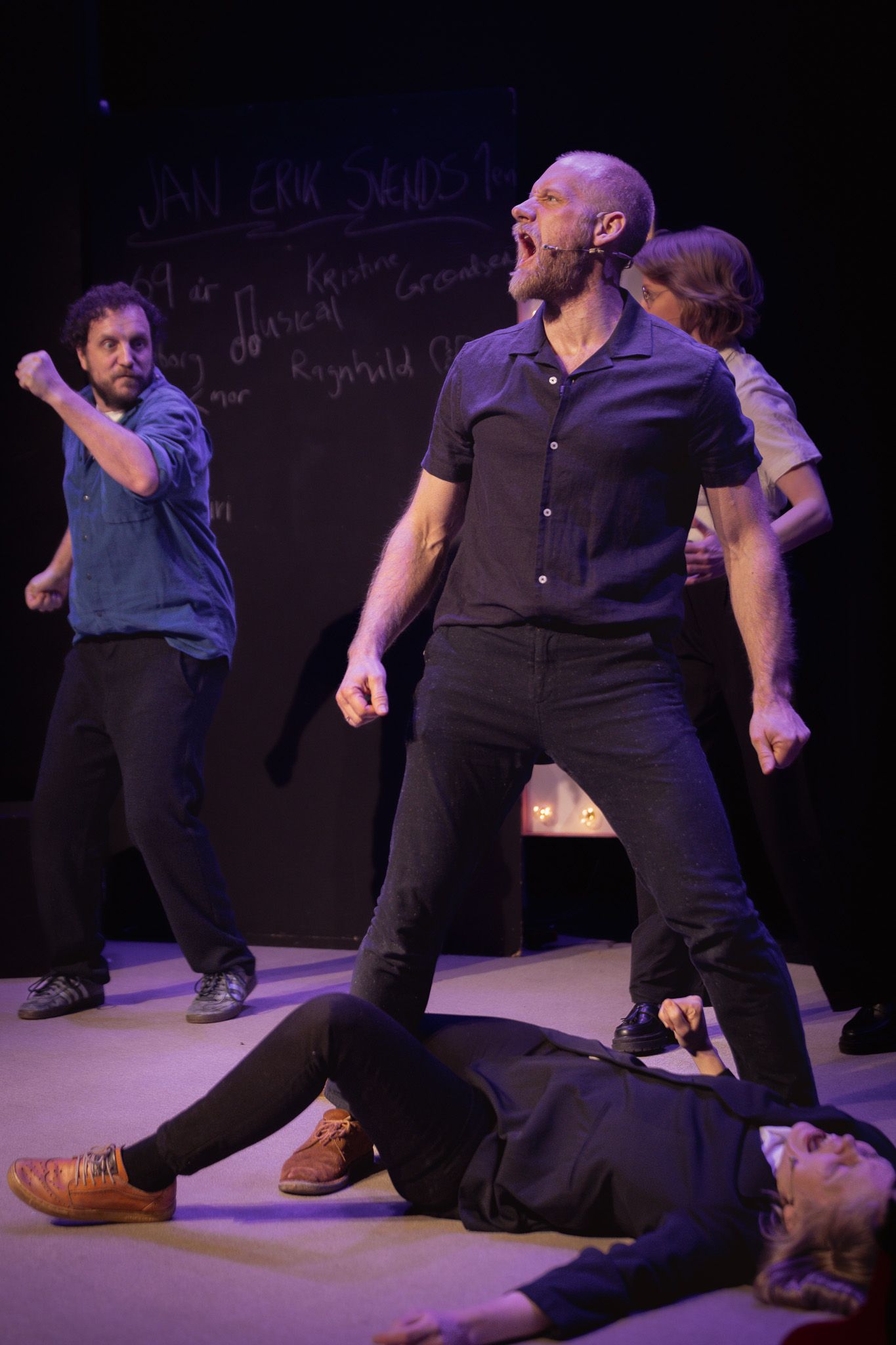 Mann i mørk skjorte står bredbeint og roper mens en person ligger på gulvet foran, andre gestikulerer bak