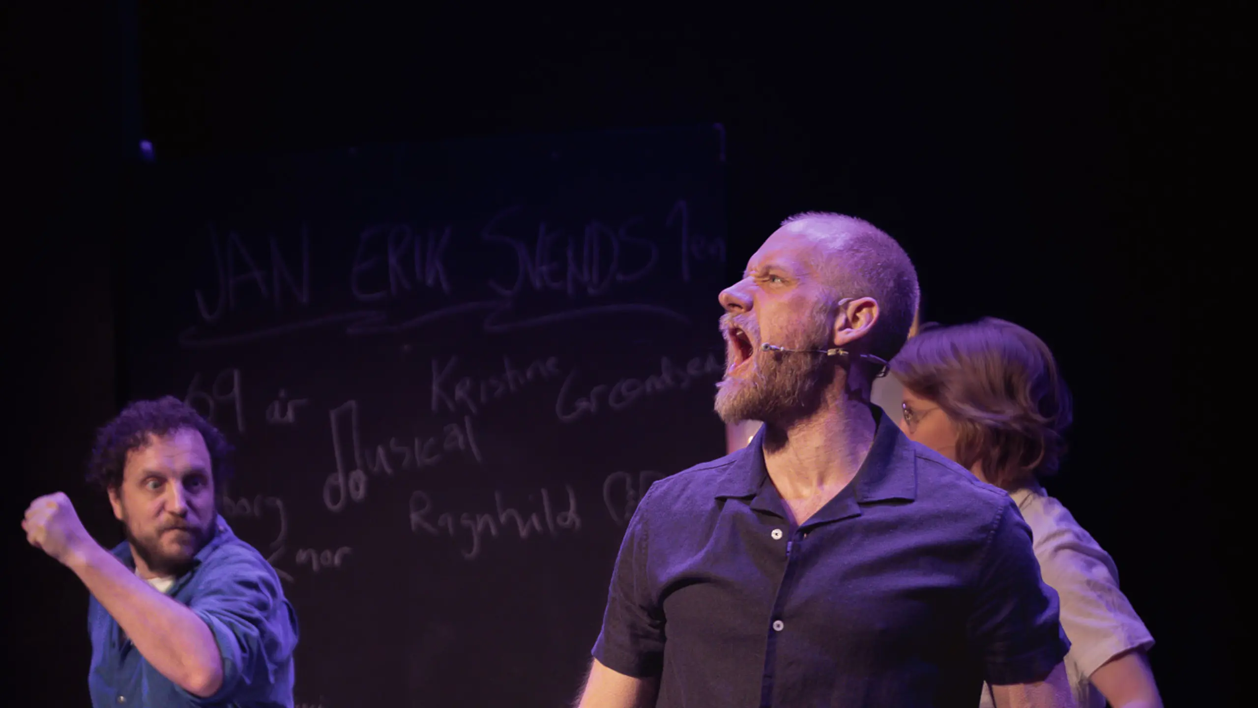 Mann i mørk skjorte står bredbeint og roper mens en person ligger på gulvet foran, andre gestikulerer bak