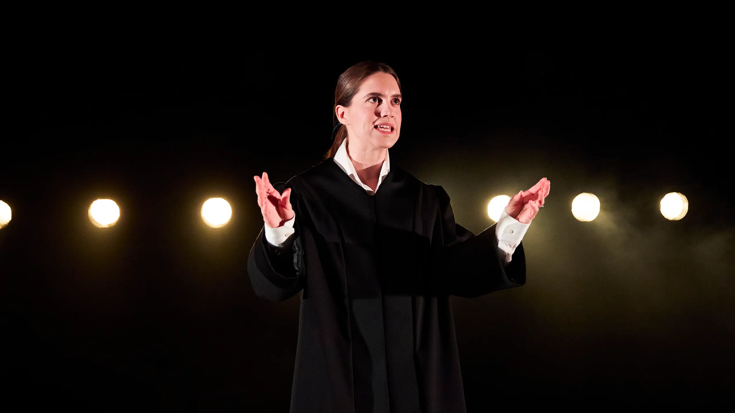 En kvinne i svart kåpe gestikulerer med hendene på en opplyst scene.
