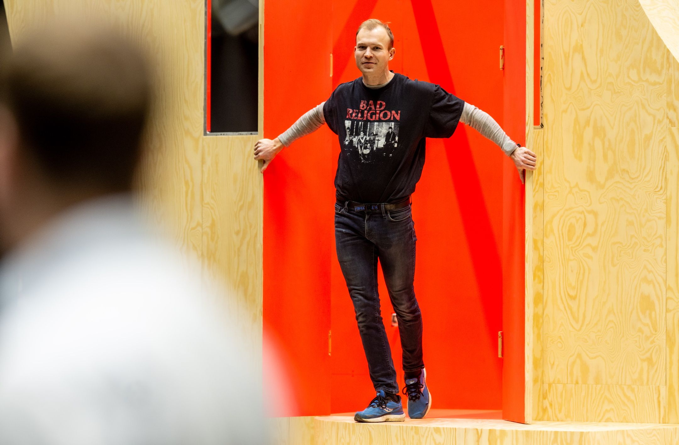 En mann med t-skjorte med bandlogo står i en døråpning med lys trefarget vegg og rød bakgrunn.
