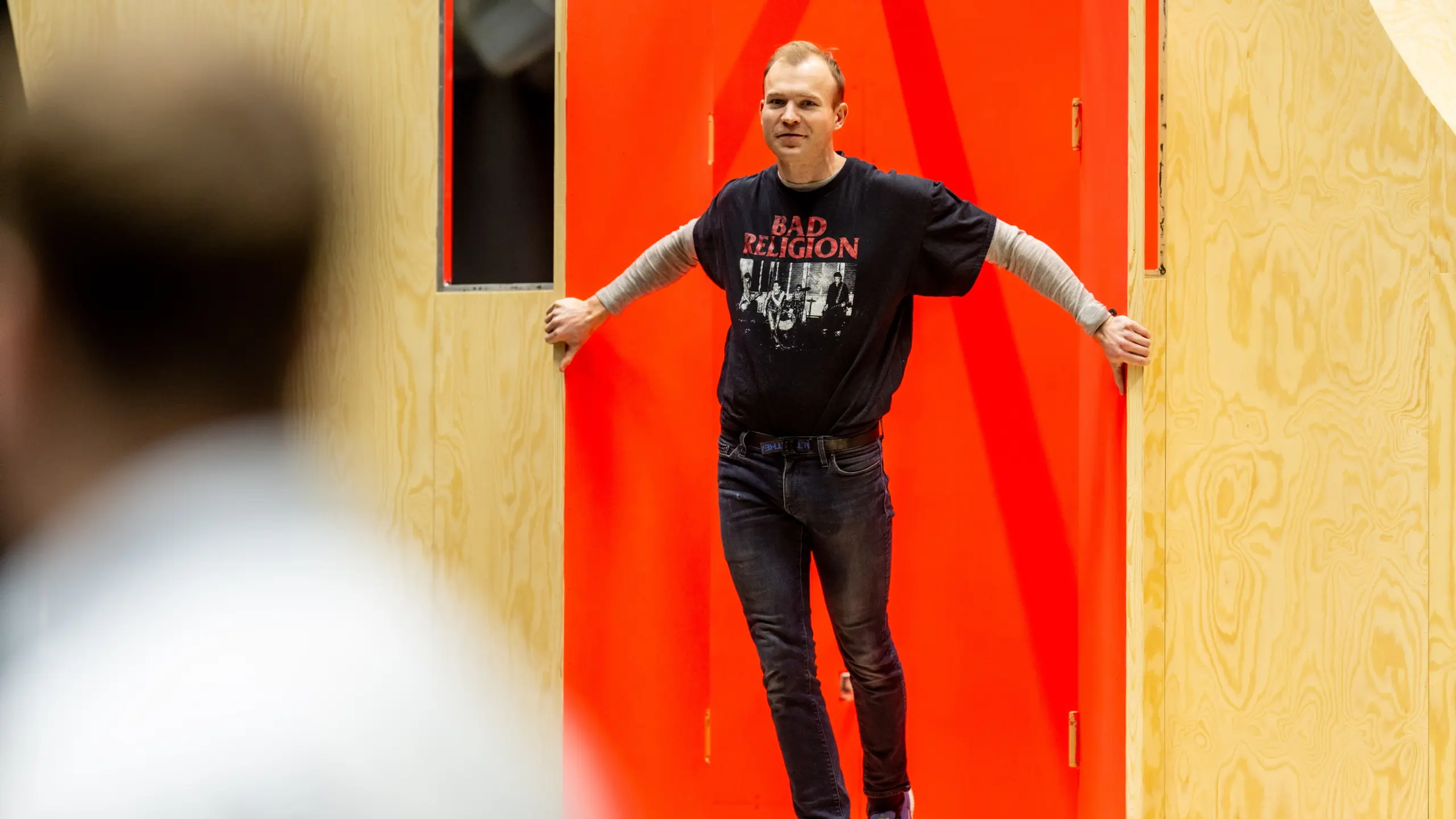 En mann med t-skjorte med bandlogo står i en døråpning med lys trefarget vegg og rød bakgrunn.