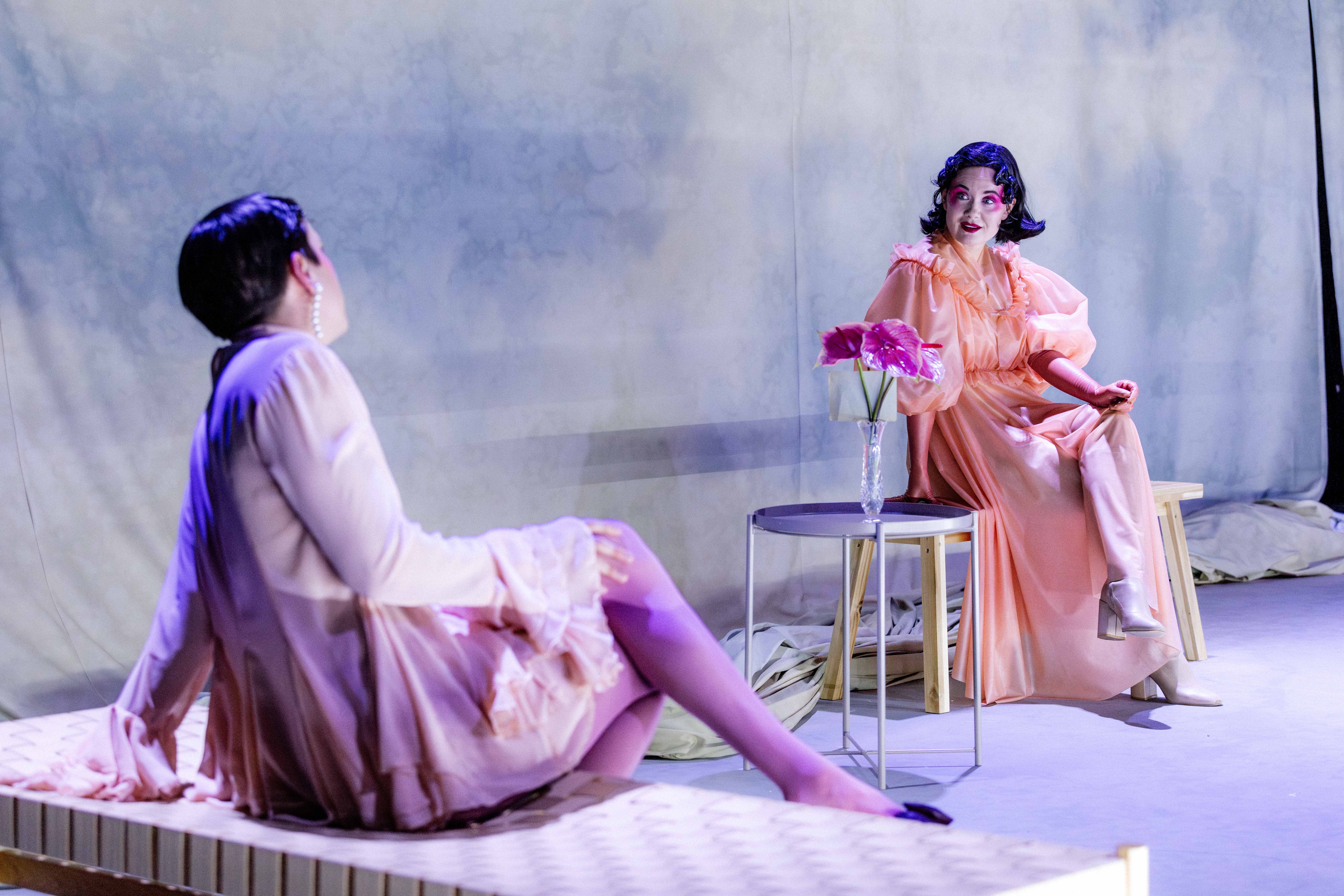 To personer sitter vendt mot hverandre på benker ved et lite bord med blomster i en vase på scenegulvet