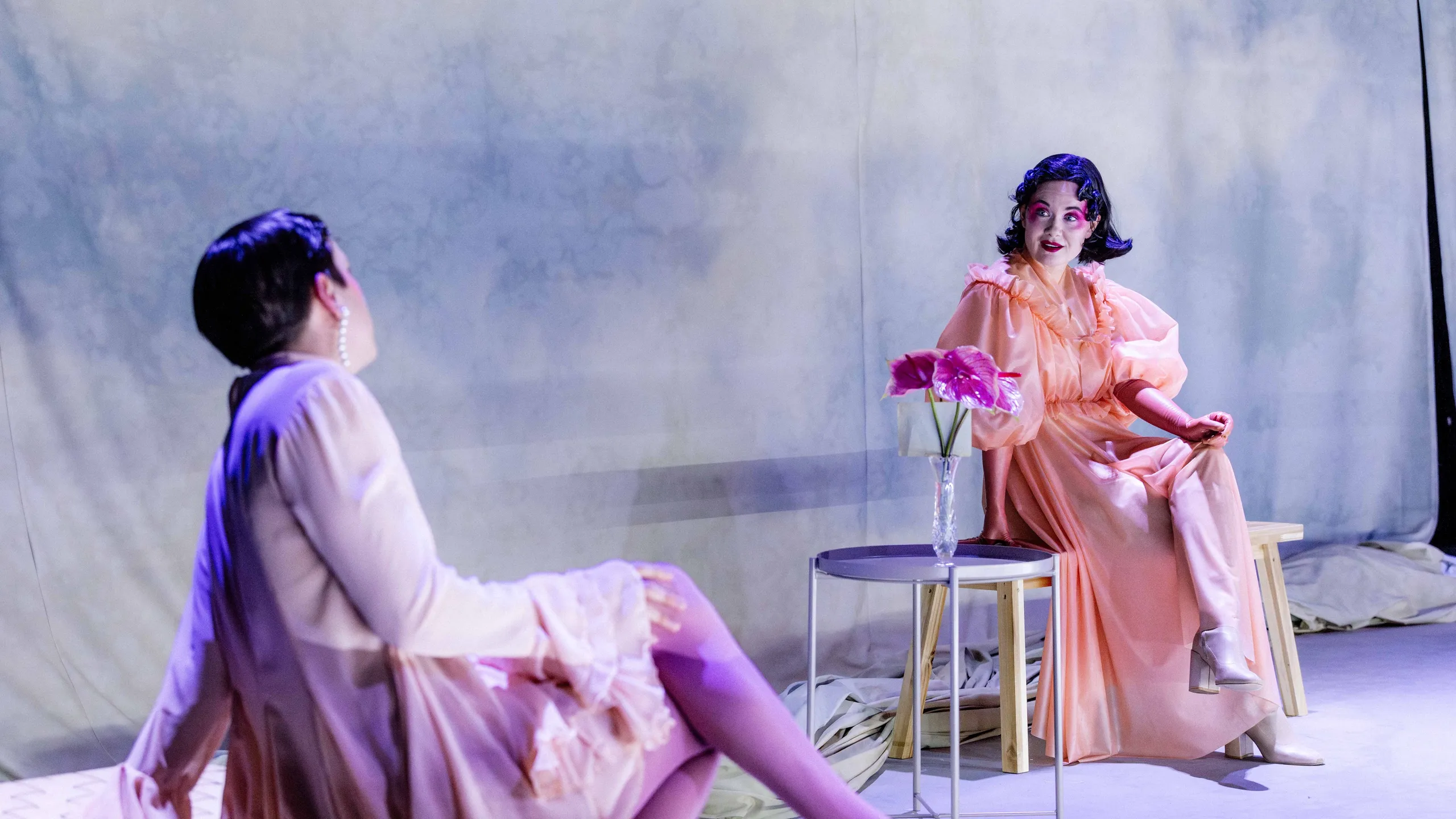 To personer sitter vendt mot hverandre på benker ved et lite bord med blomster i en vase på scenegulvet