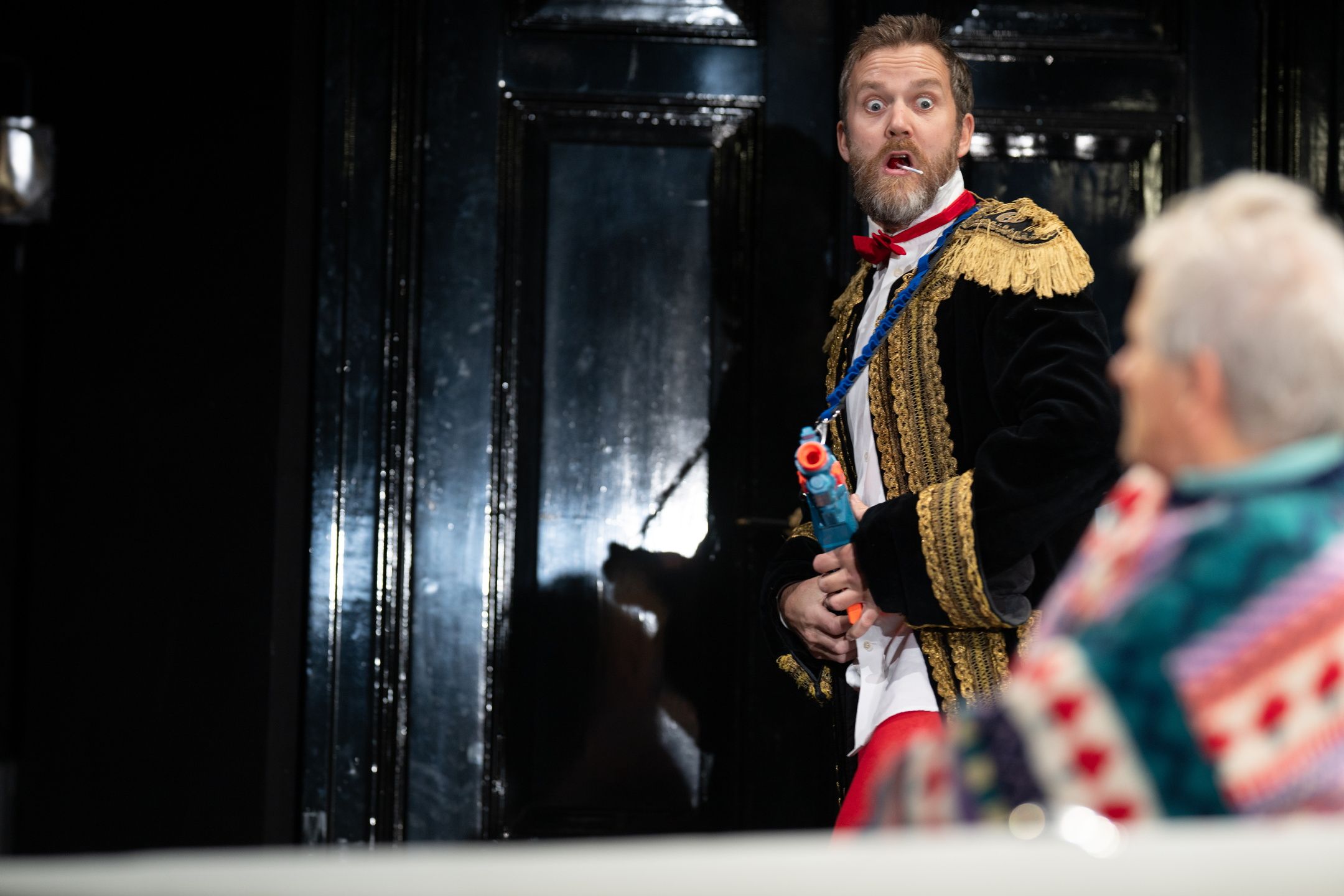 Mann i uniform med lekepistol ser overrasket ut; eldre mann i fargerik genser i forgrunnen.