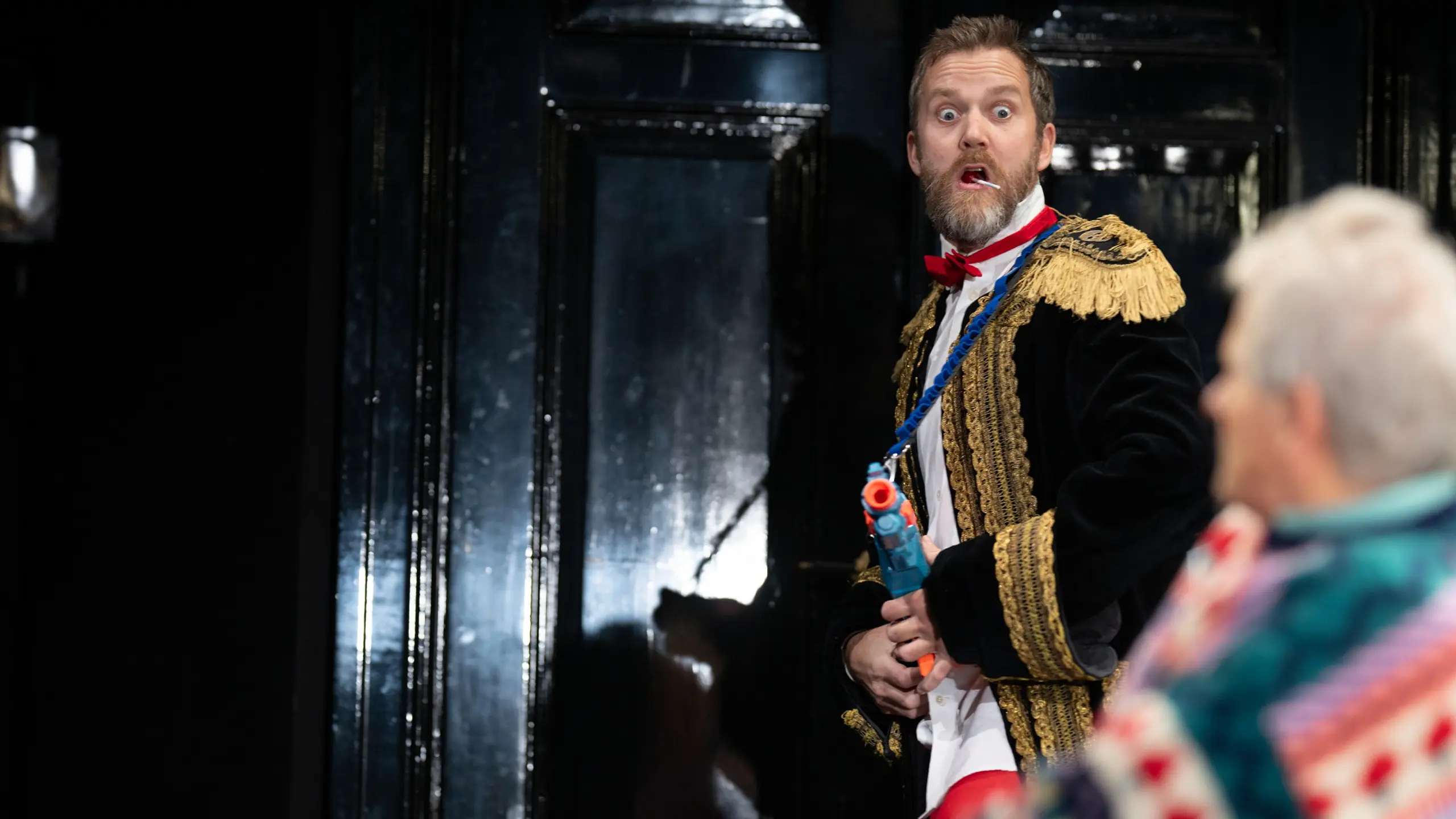 Mann i uniform med lekepistol ser overrasket ut; eldre mann i fargerik genser i forgrunnen.