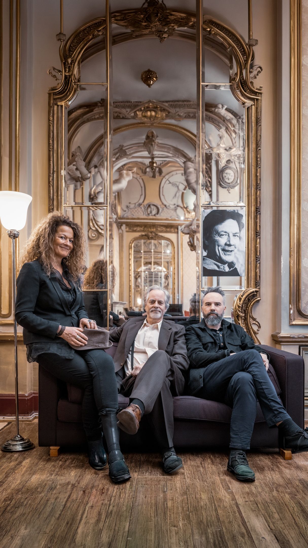 Tre mennesker sitter i en sofa i et gullforgylt rom og ser mot kameraet