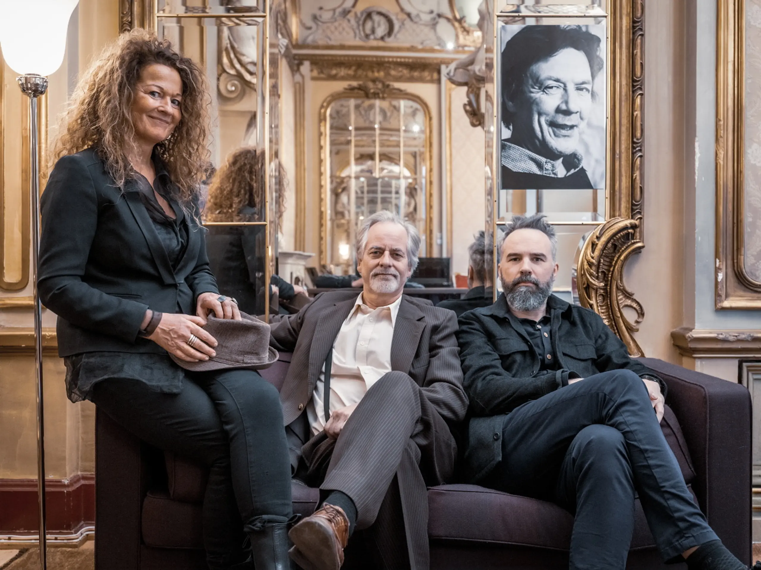 Tre mennesker sitter i en sofa i et gullforgylt rom og ser mot kameraet