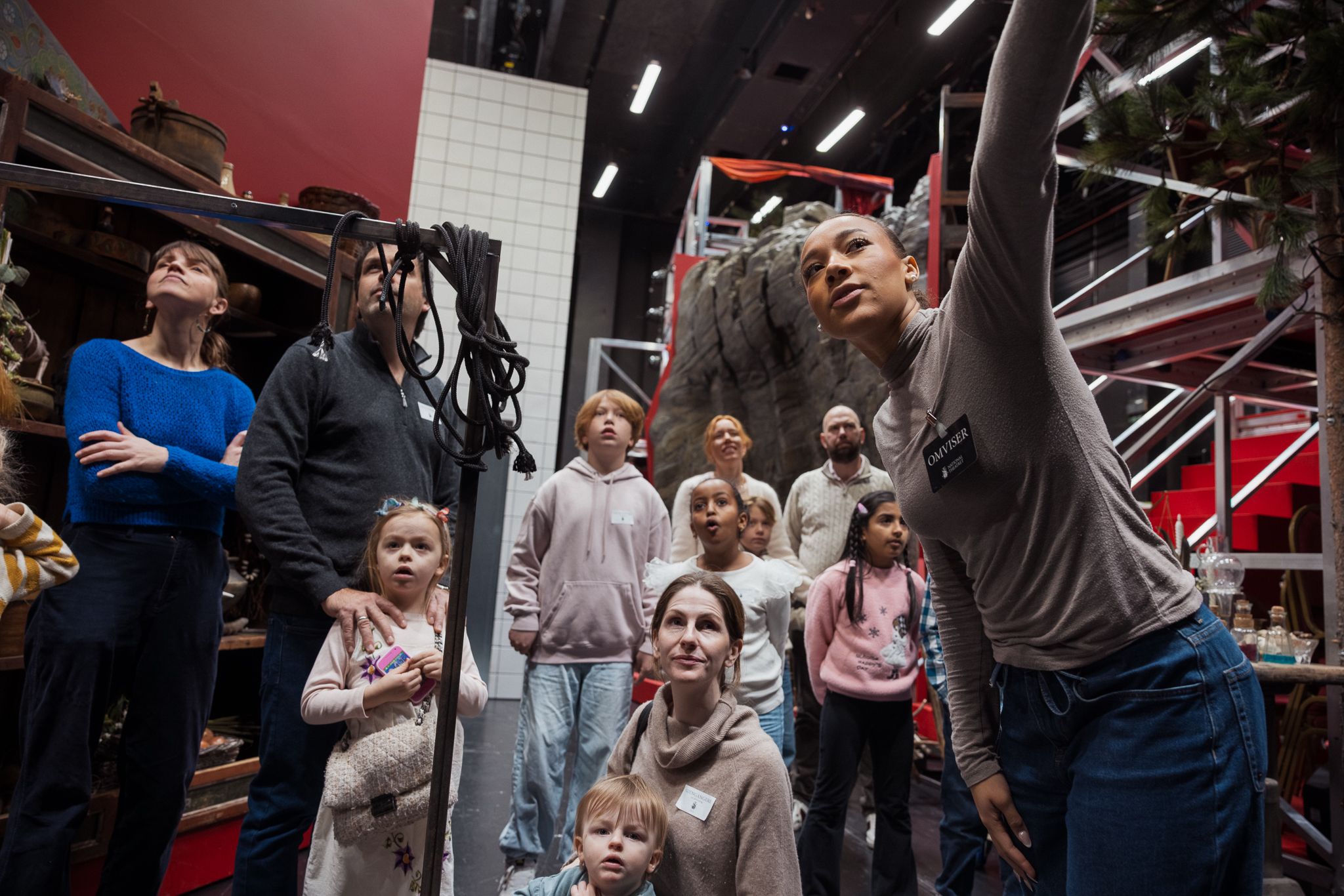 Omviser peker opp mens en gruppe barn og voksne ser på rekvisitter og kulisser i et bakrom på teateret.