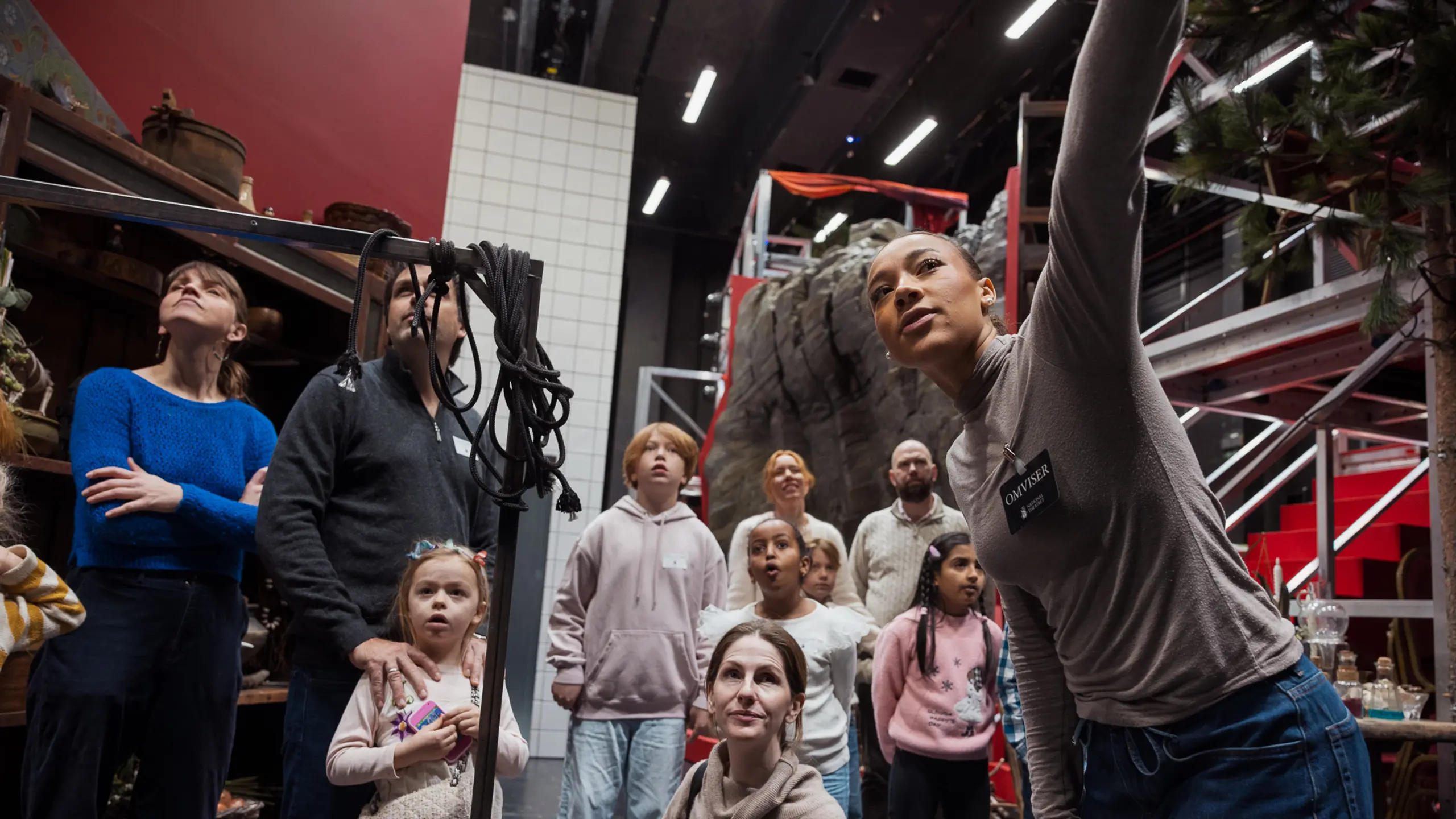 Omviser peker opp mens en gruppe barn og voksne ser på rekvisitter og kulisser i et bakrom på teateret.