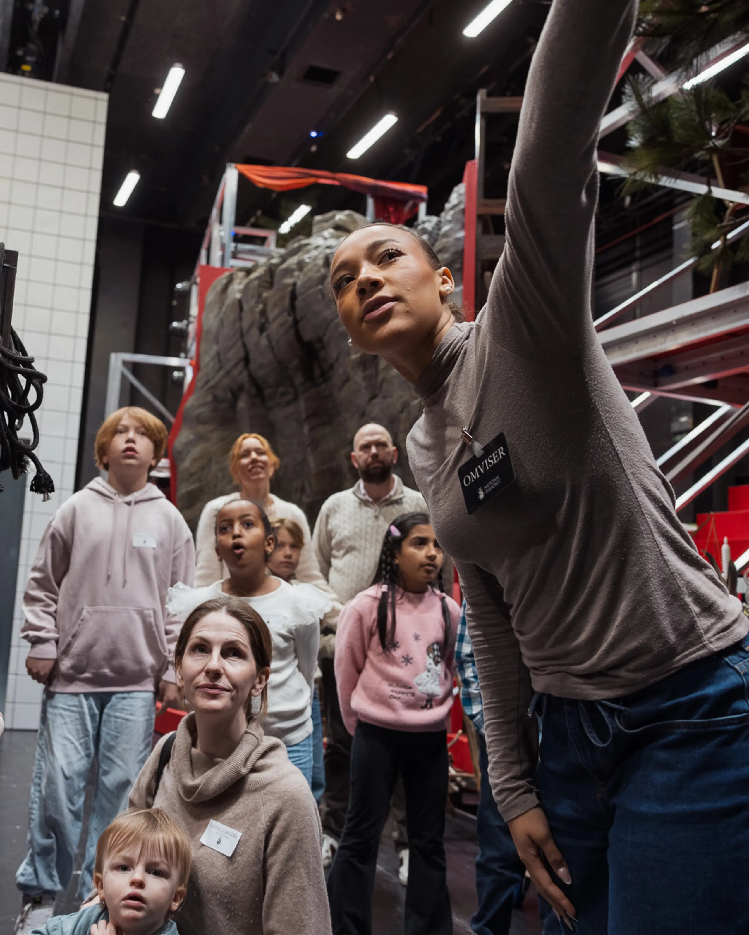 Omviser peker opp mens en gruppe barn og voksne ser på rekvisitter og kulisser i et bakrom på teateret.