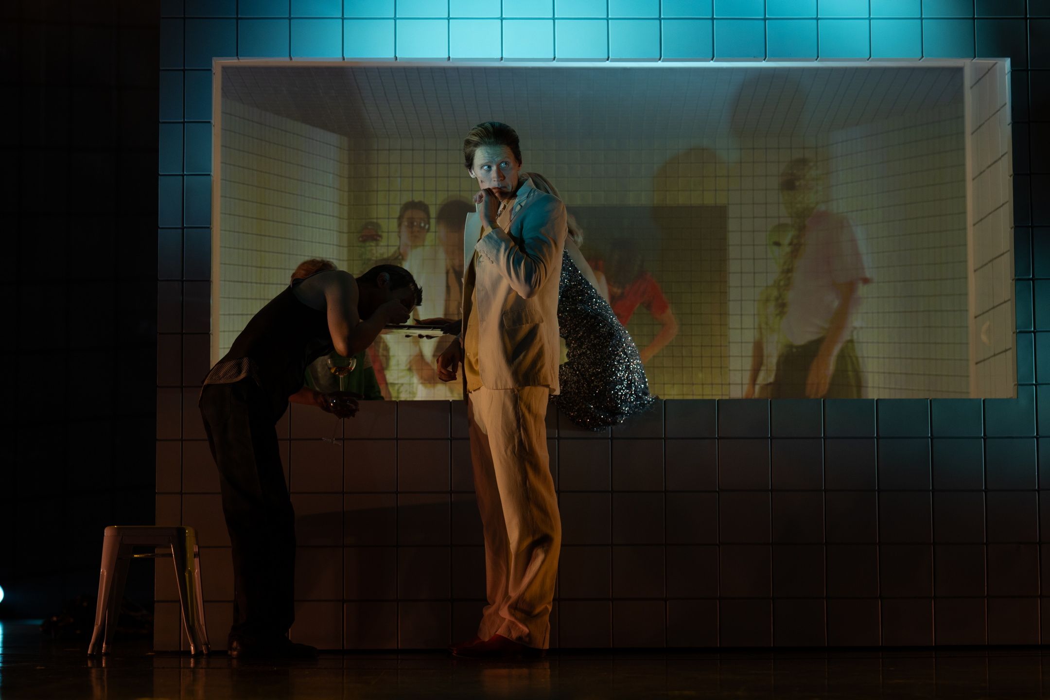 Mann i lys dress står foran flislagt vegg med innfelt videoskjerm, med skuespillere og lys bak ham.