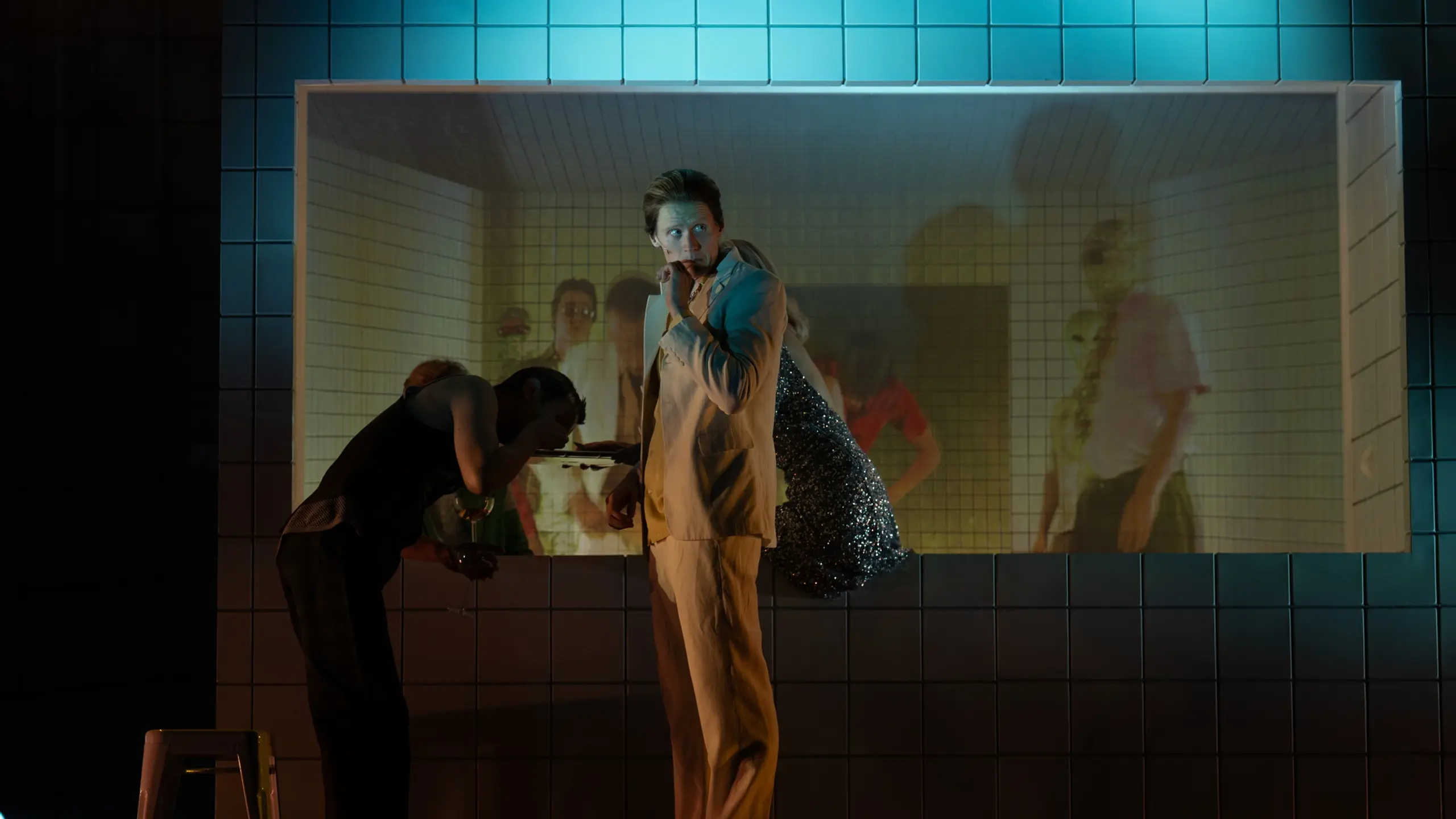 Mann i lys dress står foran flislagt vegg med innfelt videoskjerm, med skuespillere og lys bak ham.