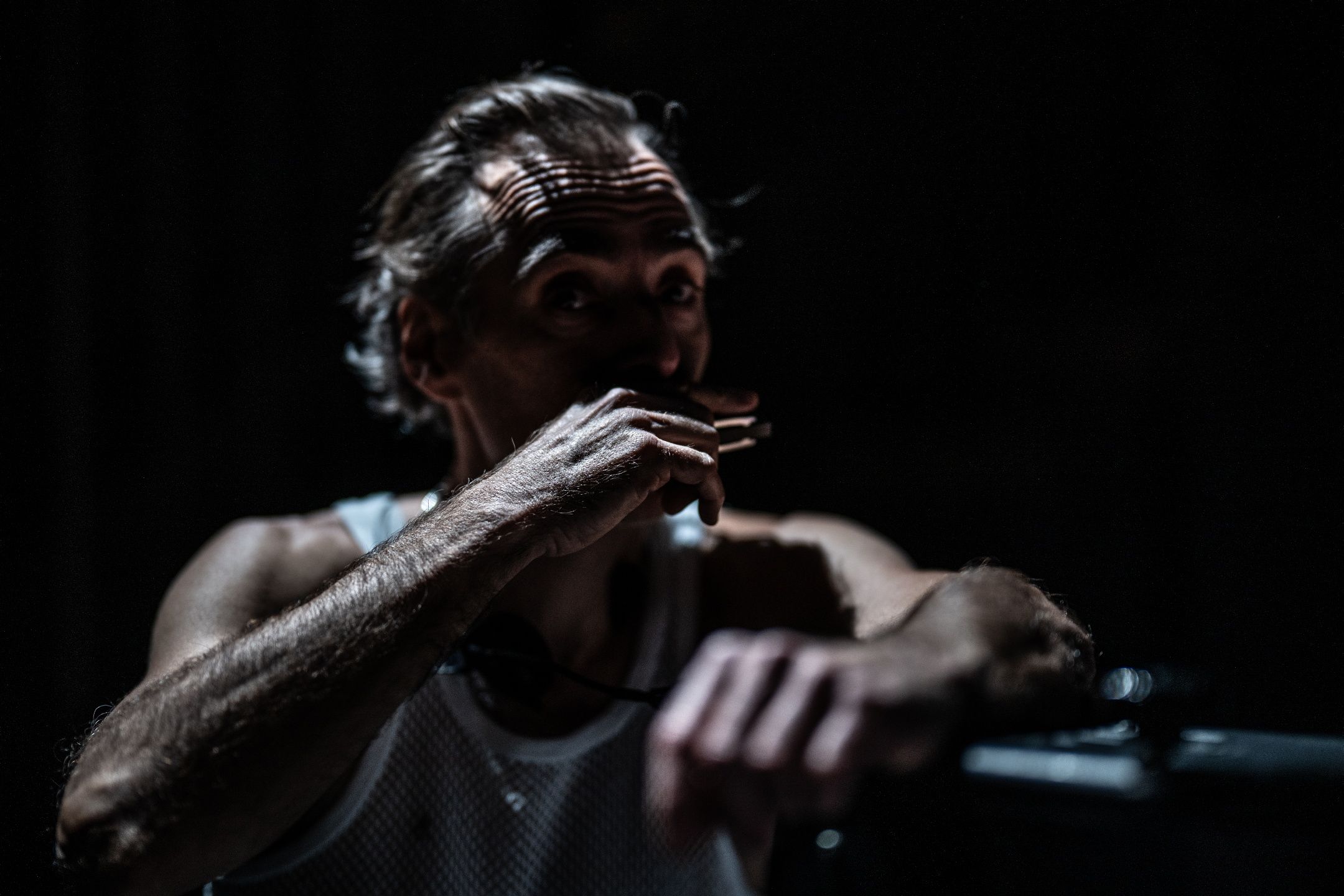 Mann i hvit nettingtrøye på mørklagt scene, dramatisk lys faller på ansikt og arm