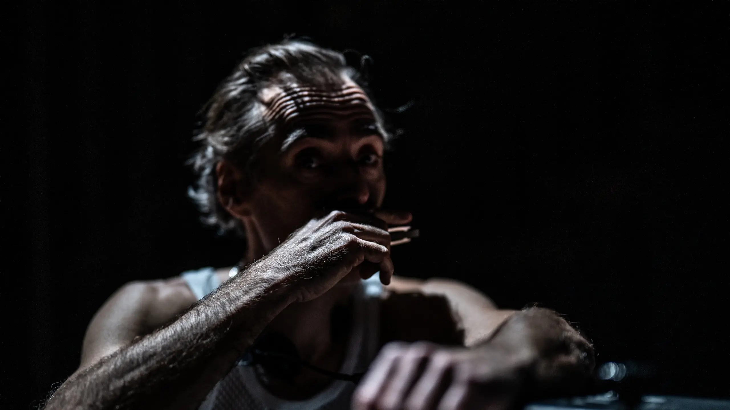 Mann i hvit nettingtrøye på mørklagt scene, dramatisk lys faller på ansikt og arm