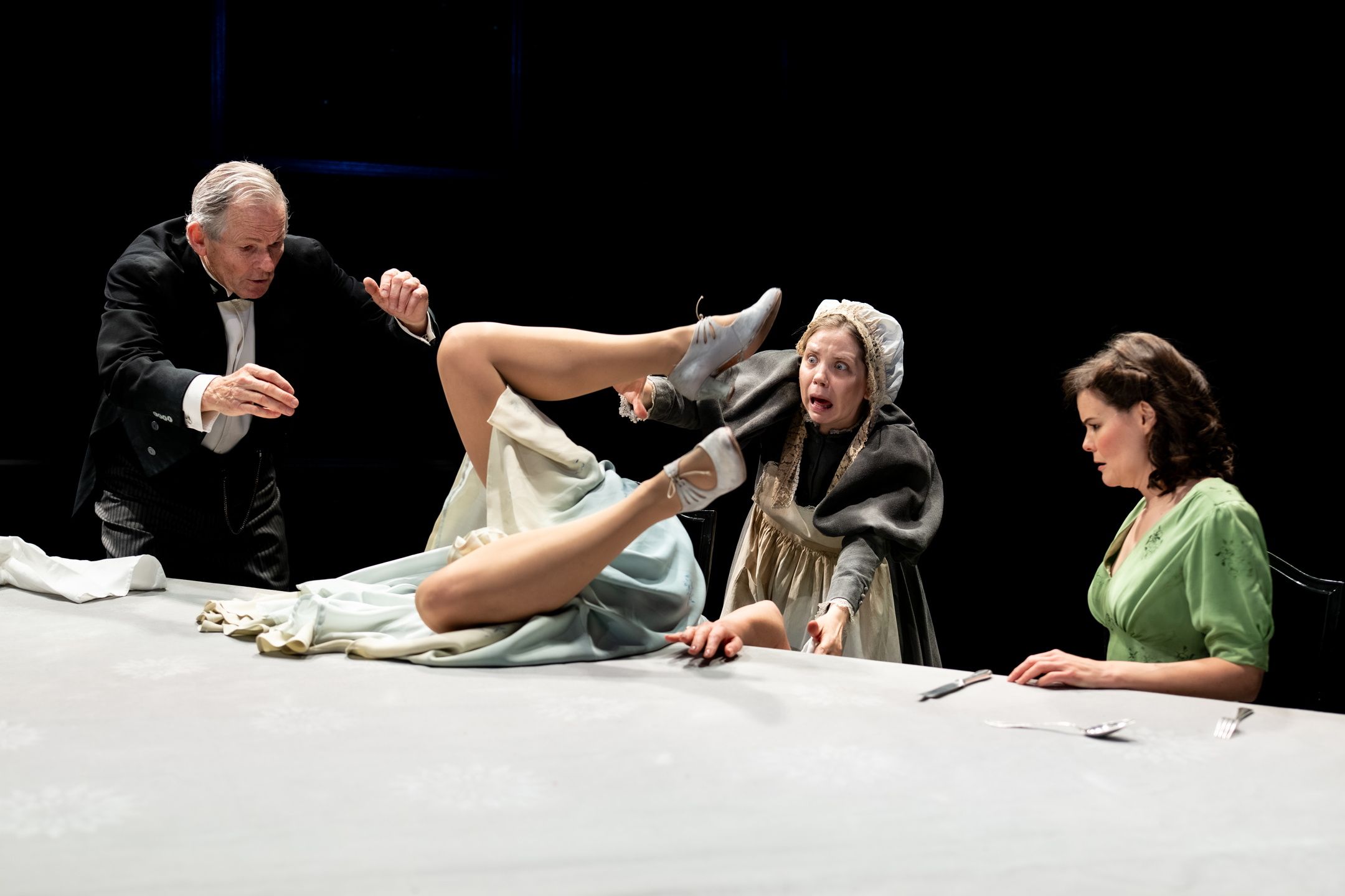 Kvinne faller bakover på bordet med beina i været, omgitt av tre personer i ulike kostymer.