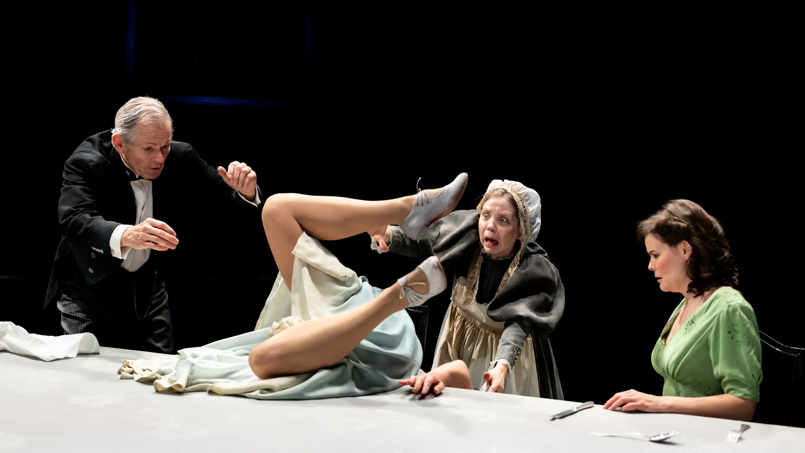 Kvinne faller bakover på bordet med beina i været, omgitt av tre personer i ulike kostymer.