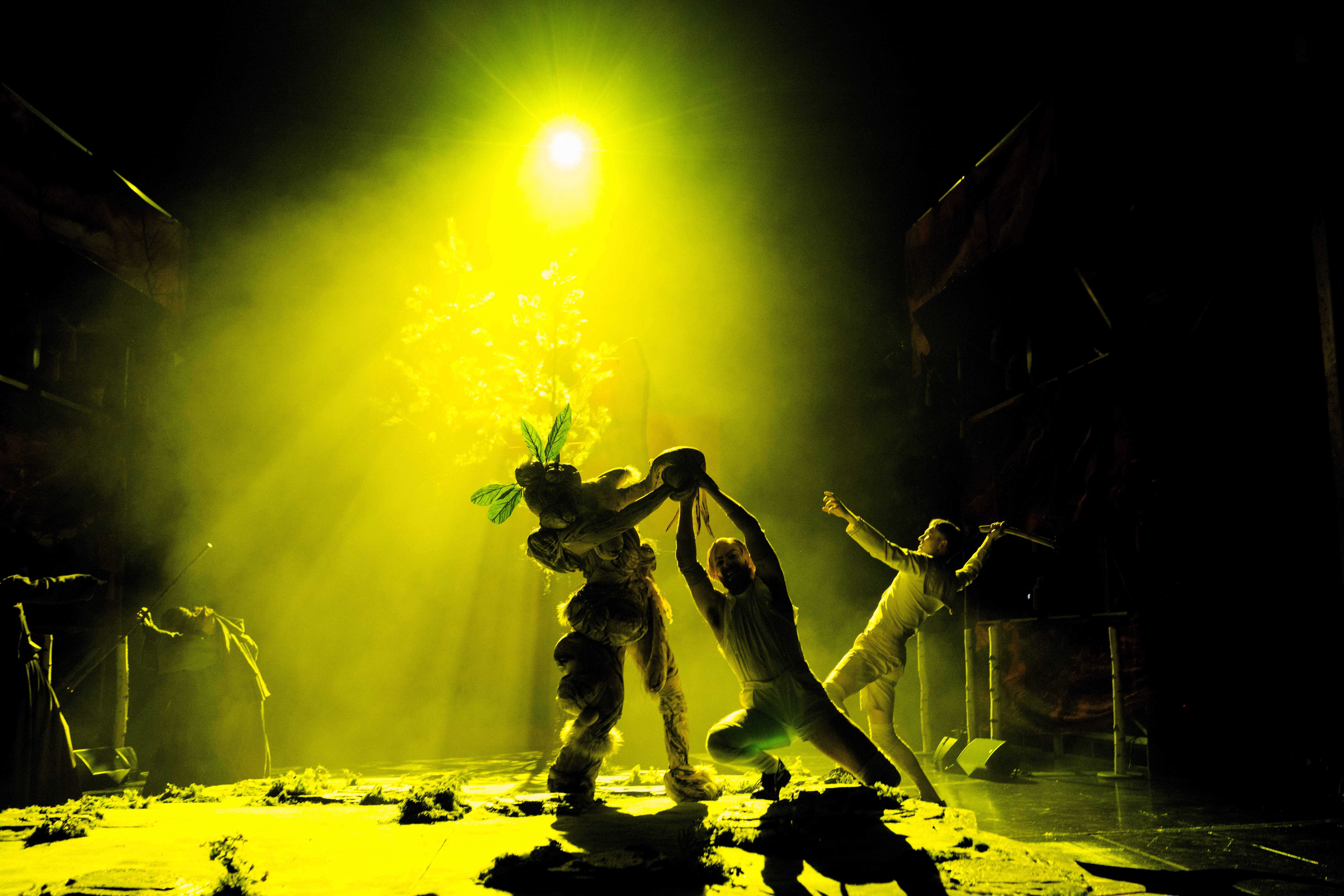 Tre utøvere under sterkt gult scenelys; en i planteaktig kostyme holder en stor rekvisitt i midten.