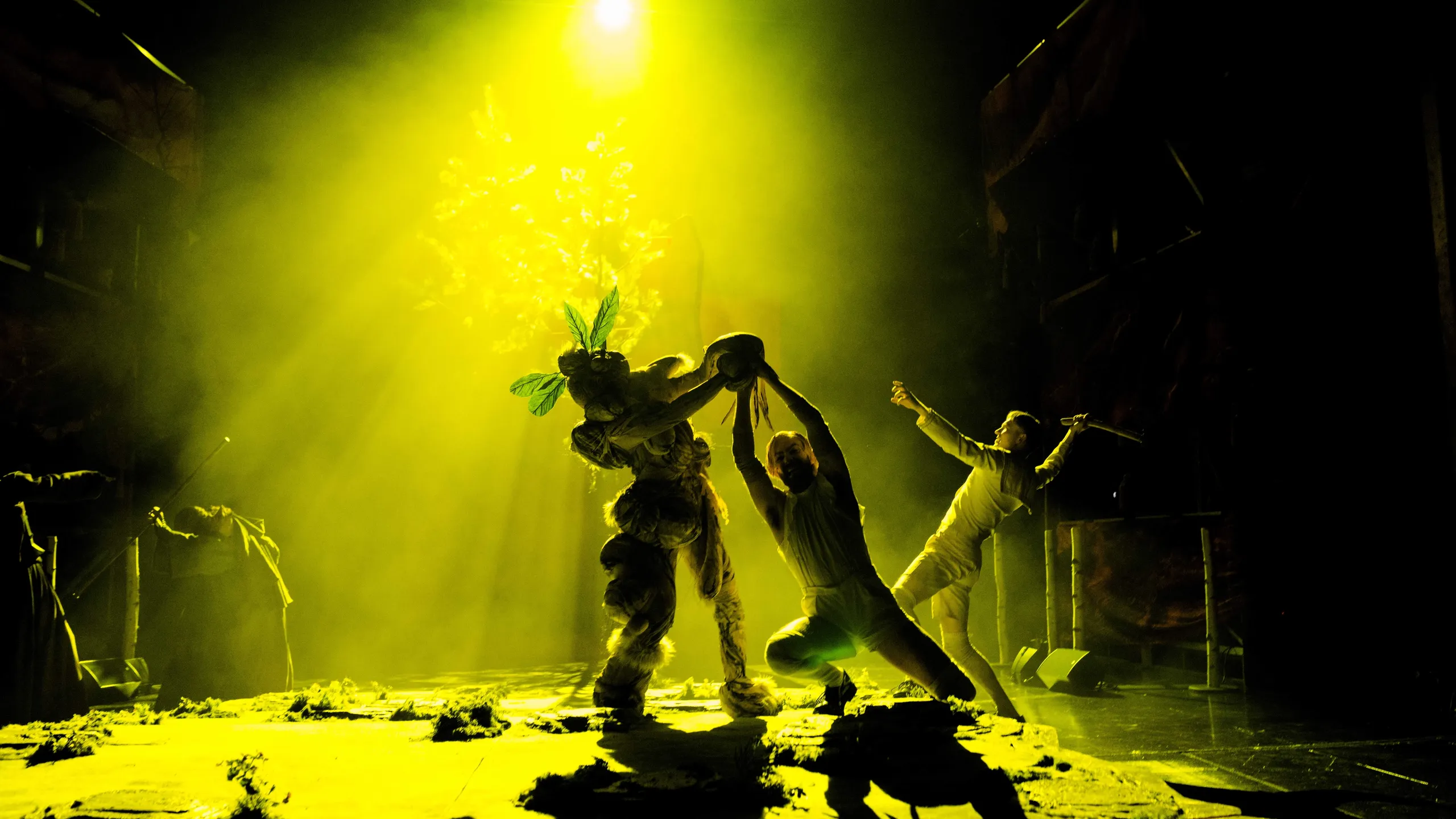Tre utøvere under sterkt gult scenelys; en i planteaktig kostyme holder en stor rekvisitt i midten.