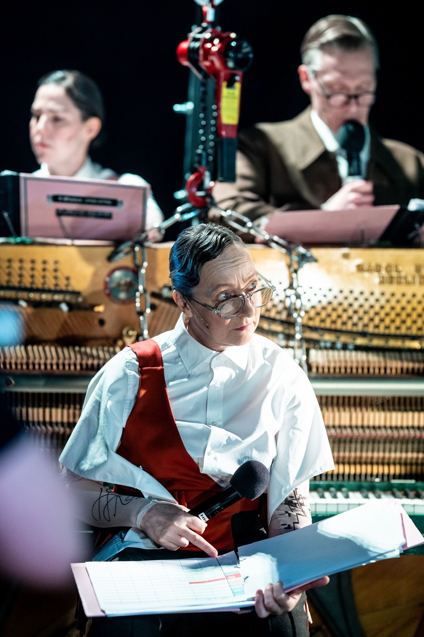Tre personer sitter ved pianoet, en av dem (i fokus) peker på et skjema og holder mikrofon. Hun har briller, kort frisyre og rødt seleforkle. Bak henne ser vi to personer til