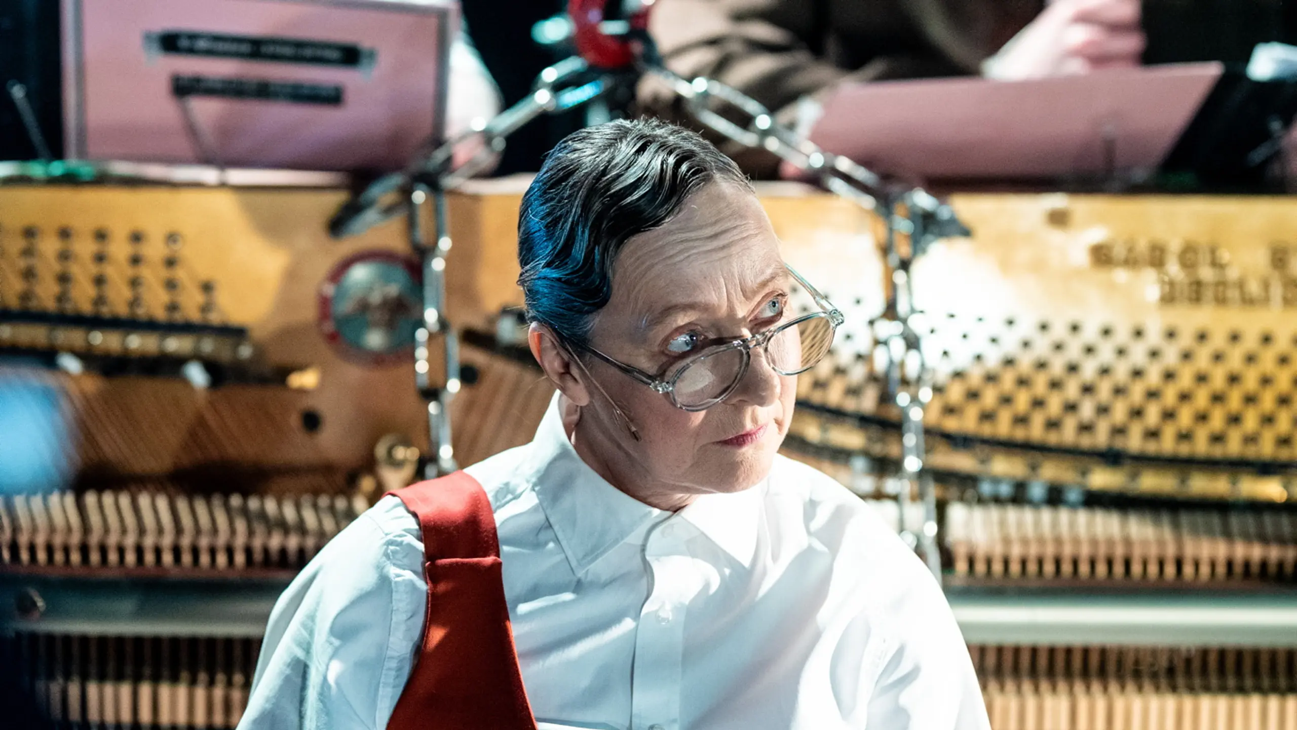 Tre personer sitter ved pianoet, en av dem (i fokus) peker på et skjema og holder mikrofon. Hun har briller, kort frisyre og rødt seleforkle. Bak henne ser vi to personer til