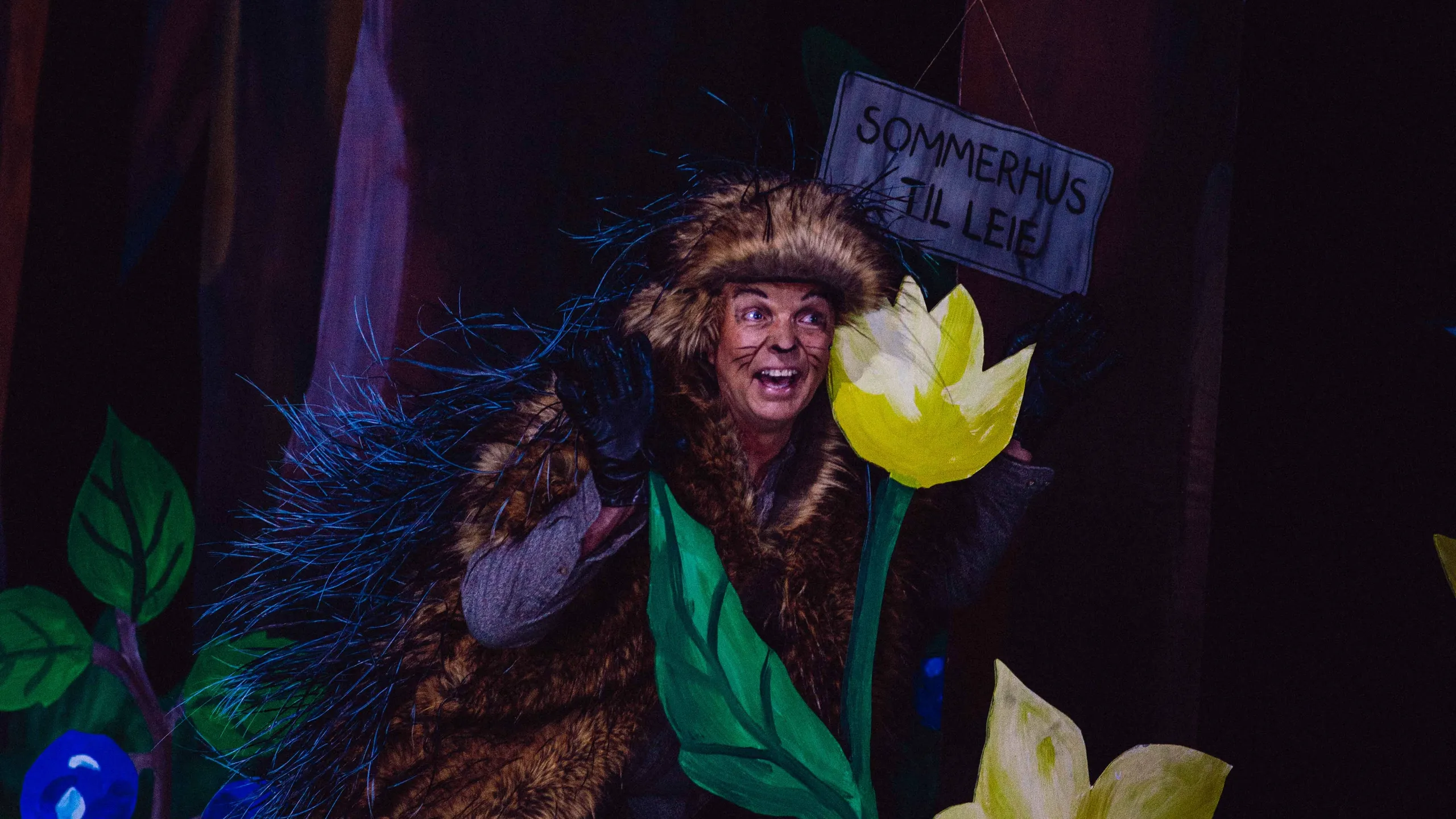 En person i pinnsvinkostyme smiler og vinker bak store blomster og et skilt med teksten 'Sommerhus til leie'