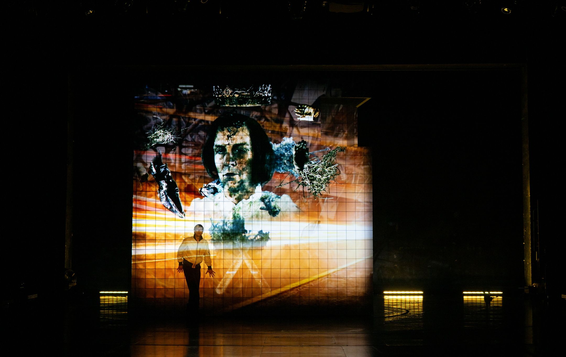 Skuespiller i hvit skjorte står foran en stor projeksjon av et ansikt med krone og mørke kontraster.