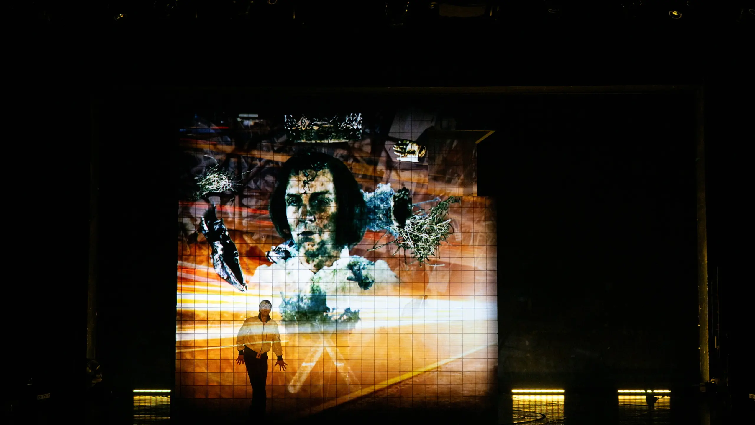 Skuespiller i hvit skjorte står foran en stor projeksjon av et ansikt med krone og mørke kontraster.