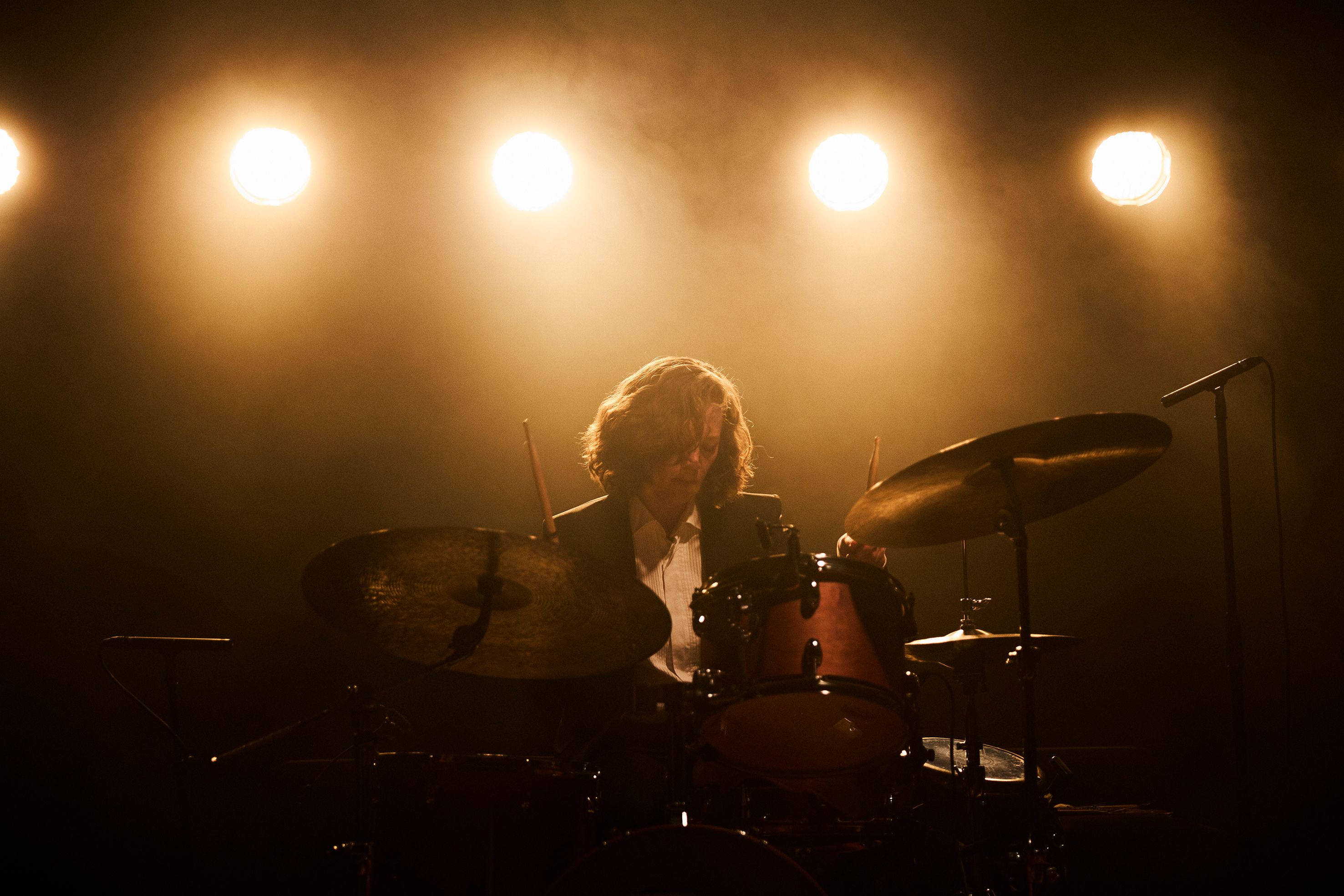En trommeslager spiller på scenen under sterke, varme lyskastere, omgitt av scenetåke.