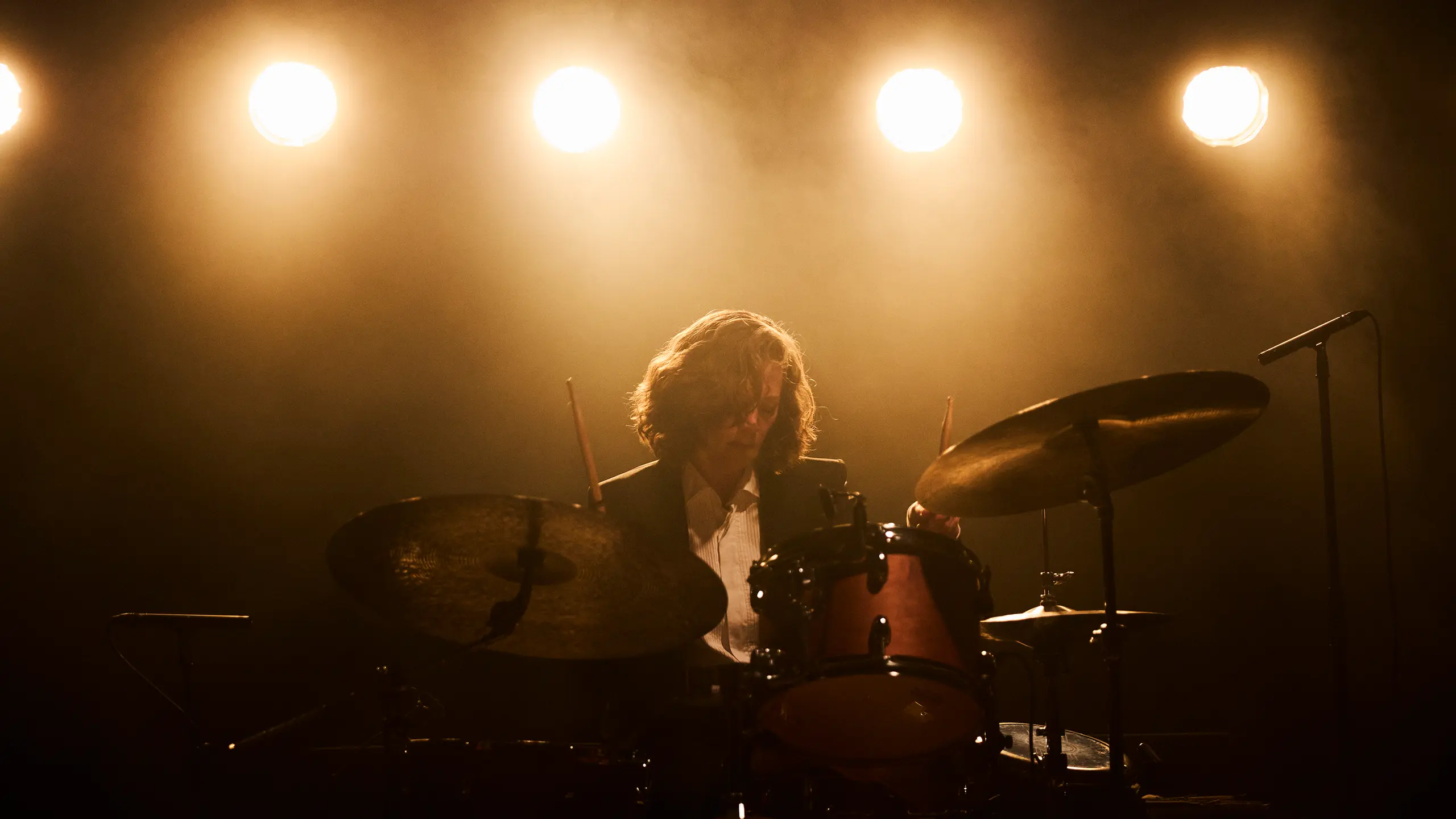 En trommeslager spiller på scenen under sterke, varme lyskastere, omgitt av scenetåke.