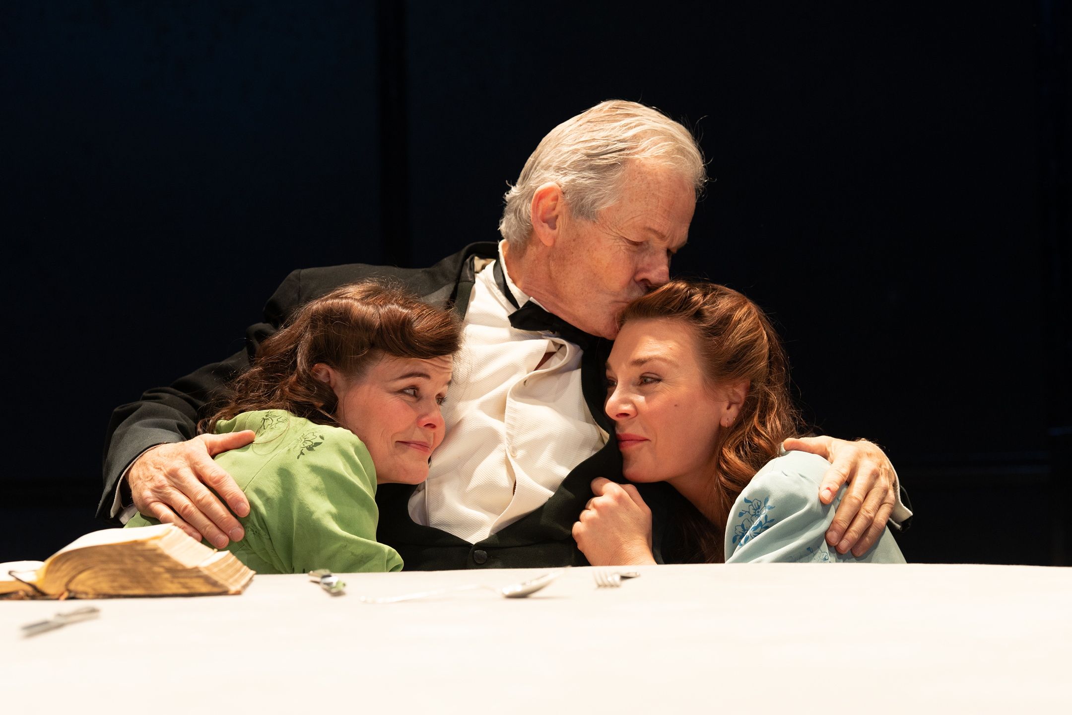 Mann i kjole og hvitt holder to kvinner inntil seg sittende ved et bord