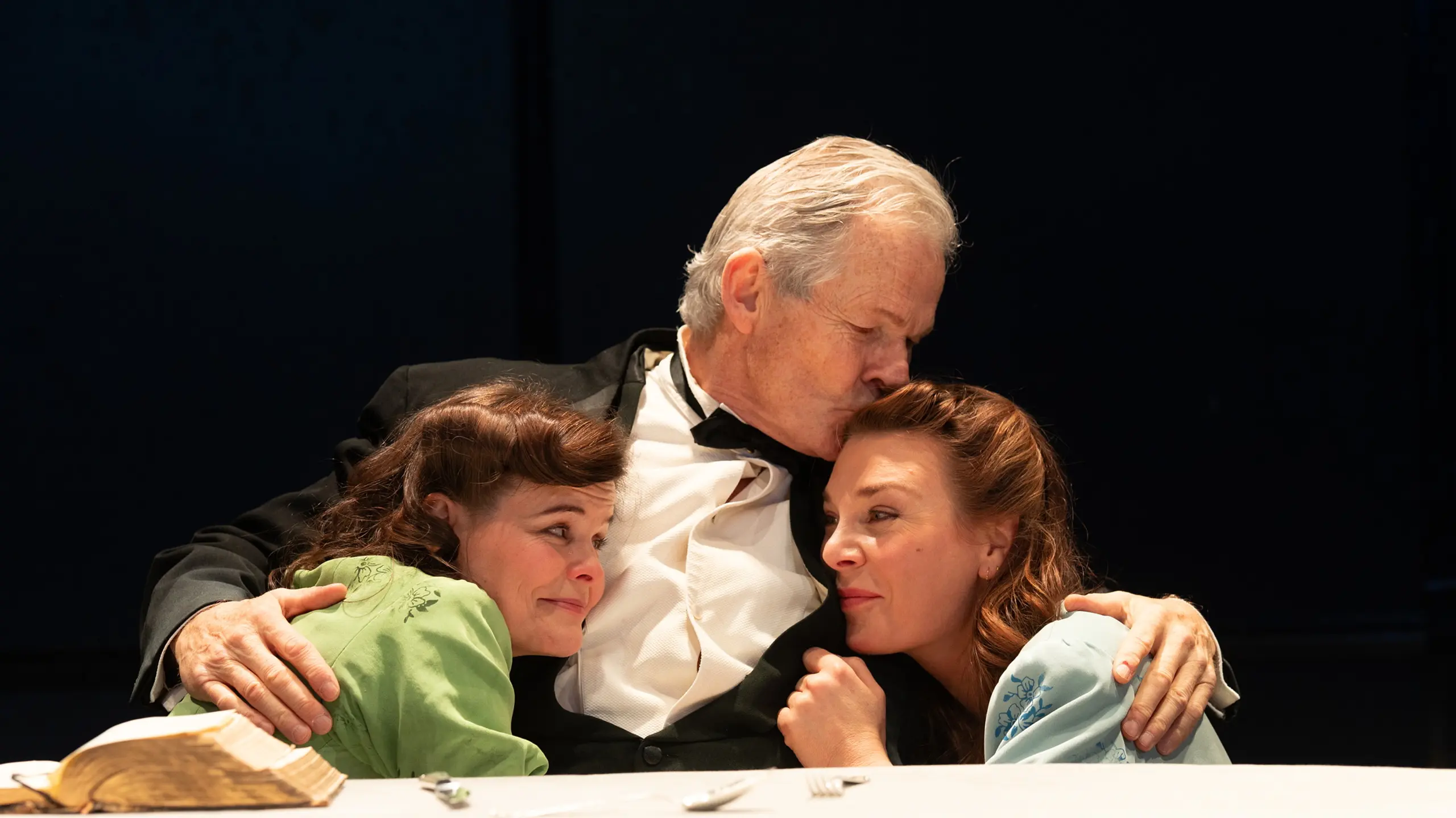 Mann i kjole og hvitt holder to kvinner inntil seg sittende ved et bord