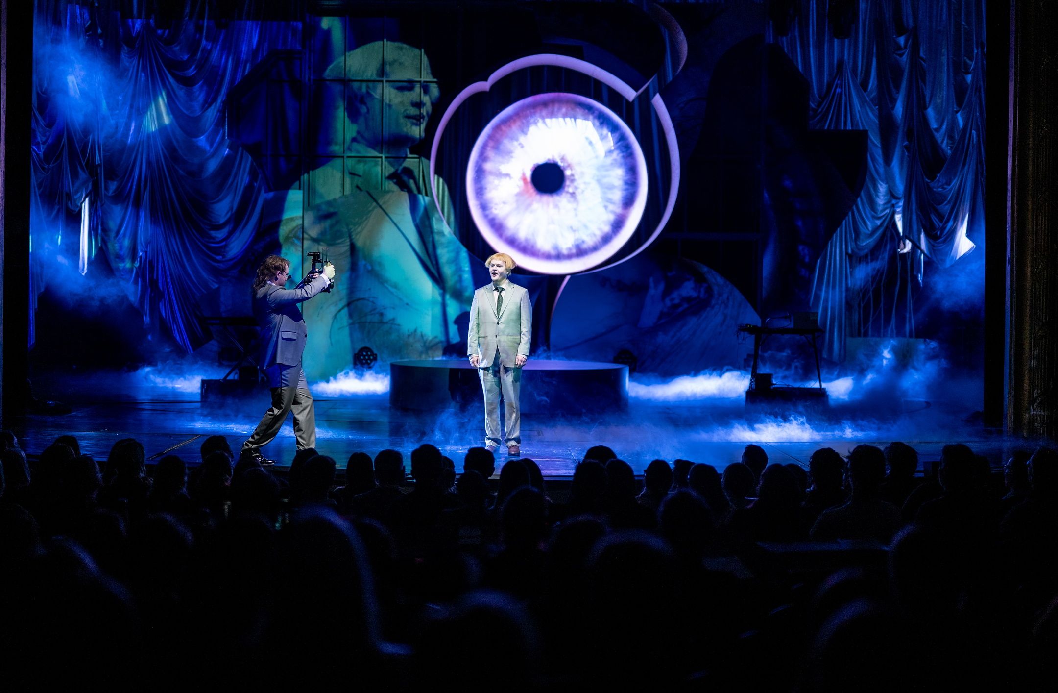 Publikum ser på to personer på scenen foran stort projisert øye og blå røyk, én blir filmet av den andre.