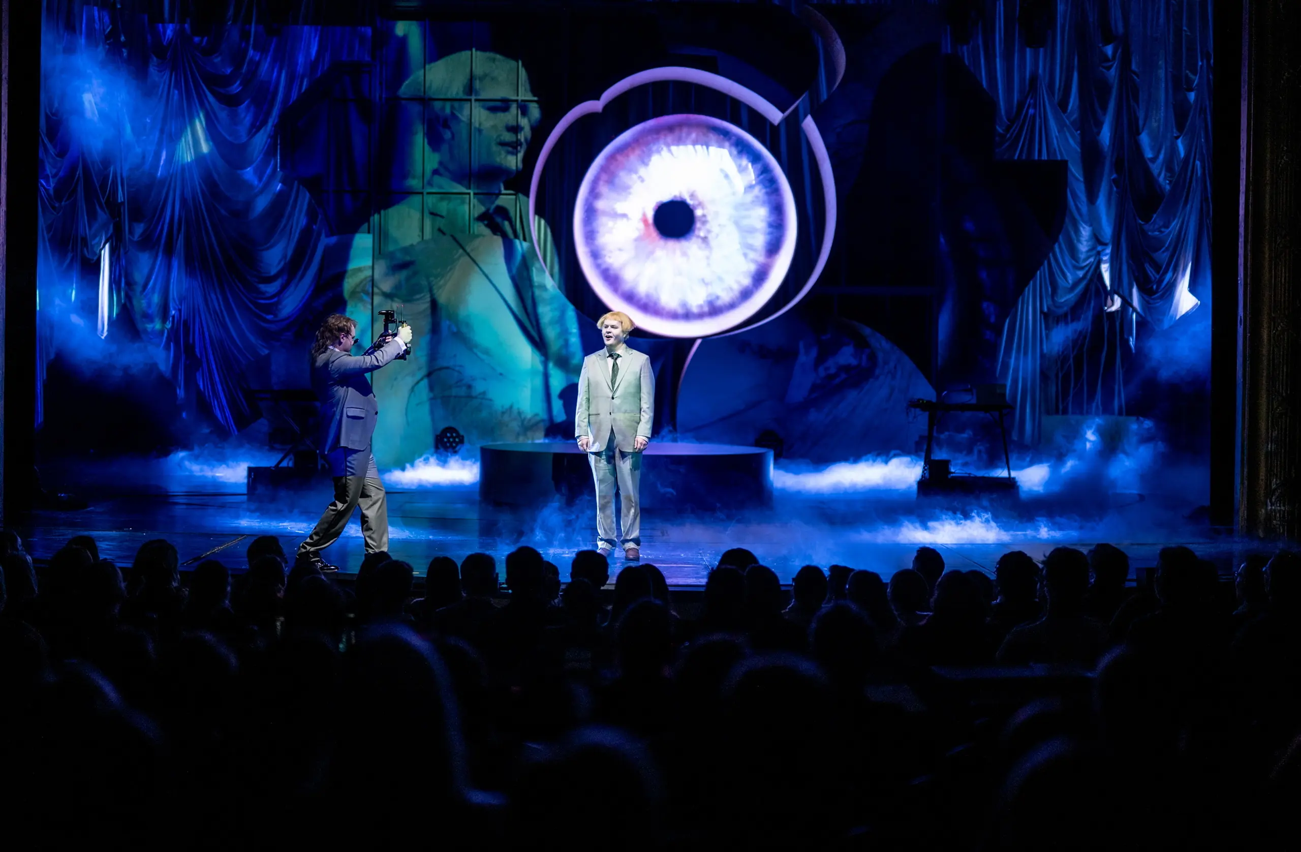 Publikum ser på to personer på scenen foran stort projisert øye og blå røyk, én blir filmet av den andre.