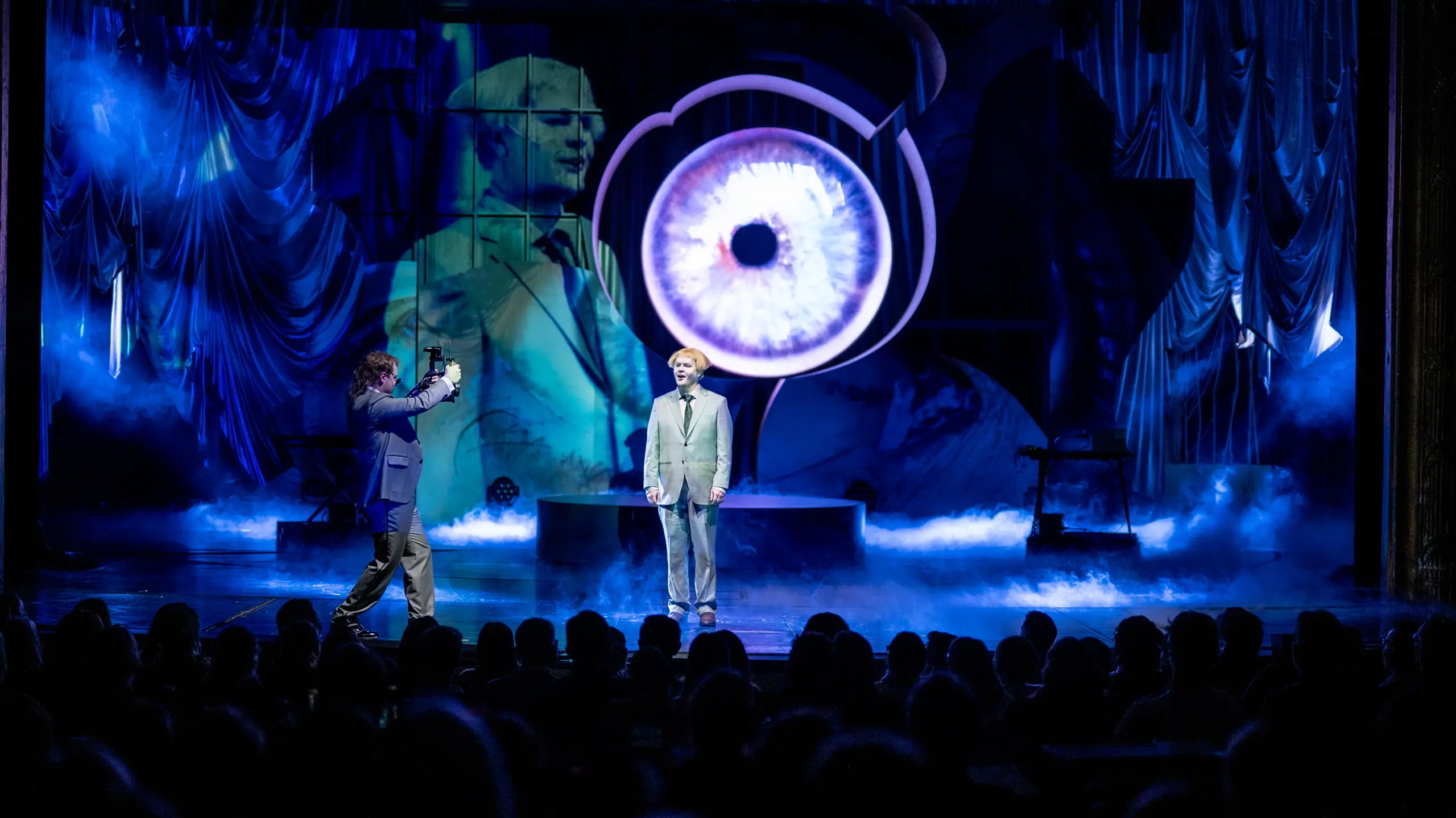 Publikum ser på to personer på scenen foran stort projisert øye og blå røyk, én blir filmet av den andre.