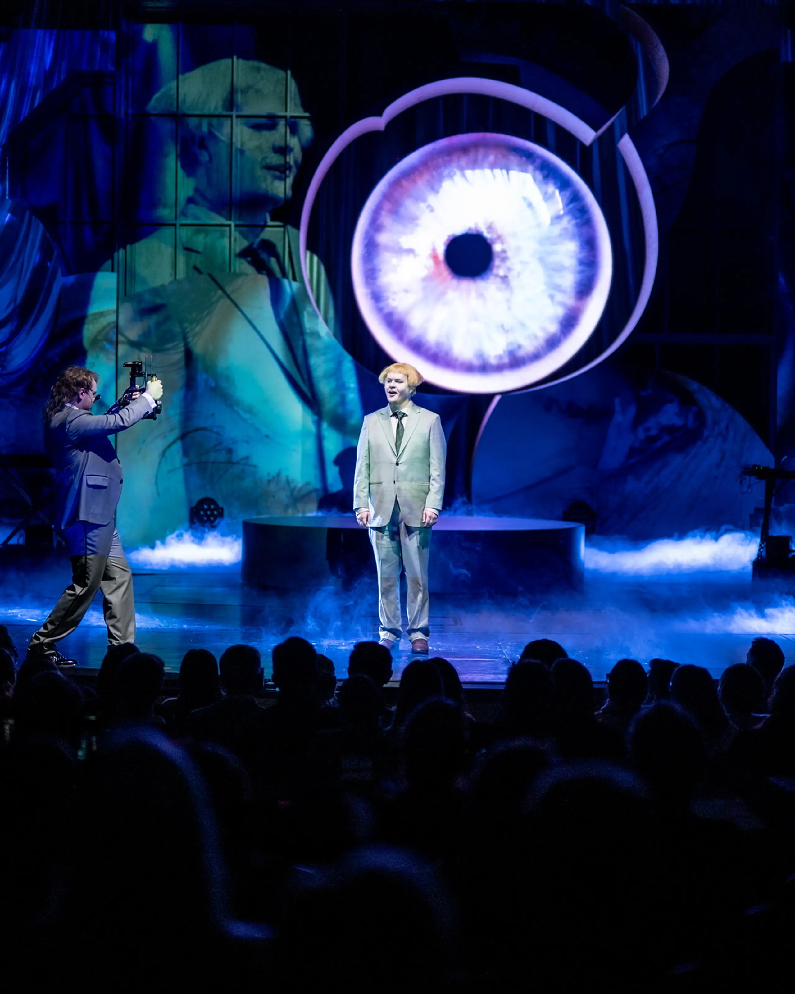 Publikum ser på to personer på scenen foran stort projisert øye og blå røyk, én blir filmet av den andre.