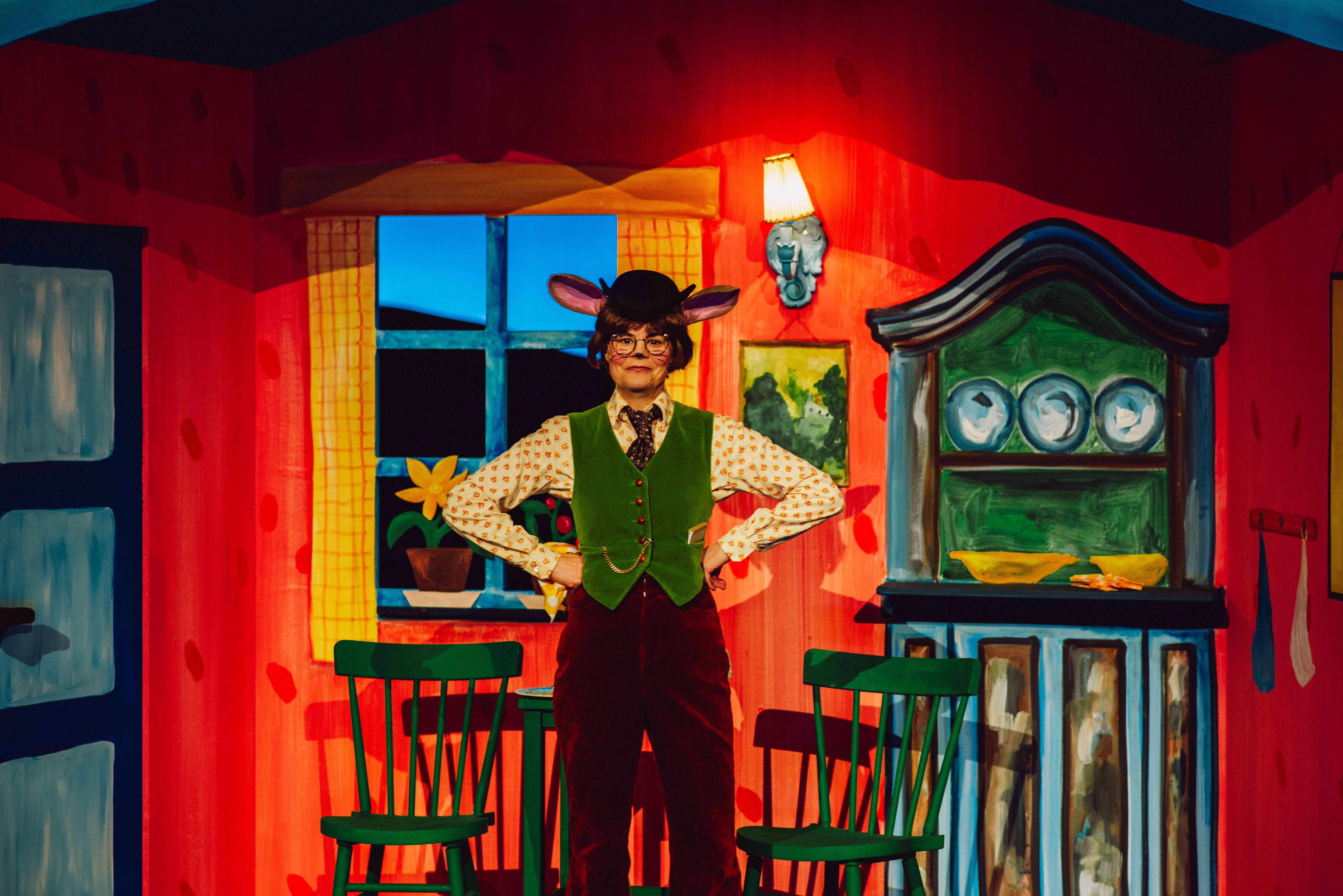 En person med museører og hatt står med hendene i siden i et fargerikt kjøkkensett med grønne stoler og servise.
