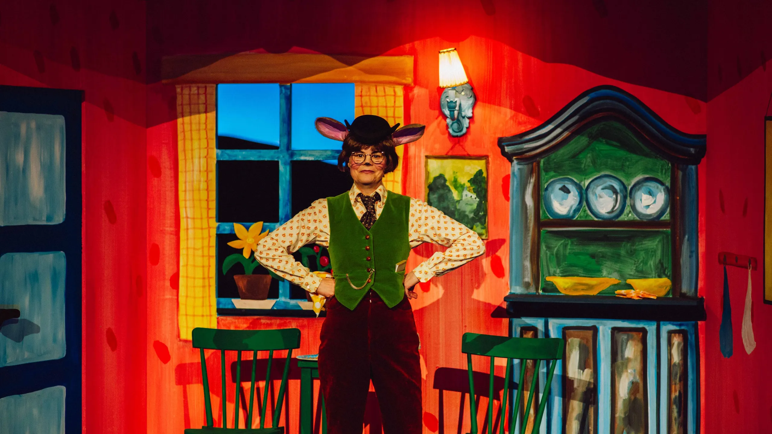 En person med museører og hatt står med hendene i siden i et fargerikt kjøkkensett med grønne stoler og servise.