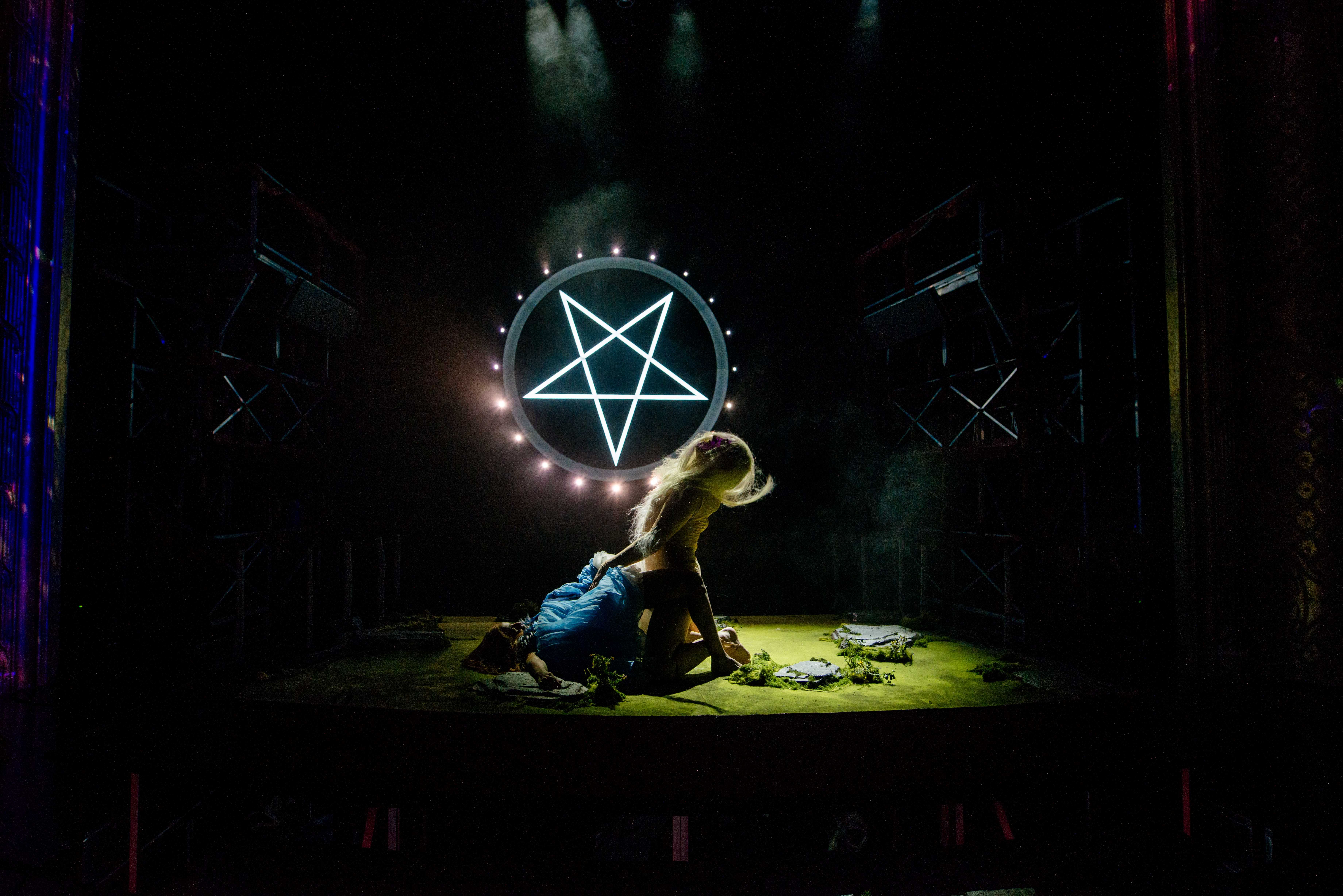 Person med langt lyst hår sitter over en annen i blå kjole foran en lysende pentagramring, i mørk og tåkete teaterscene.