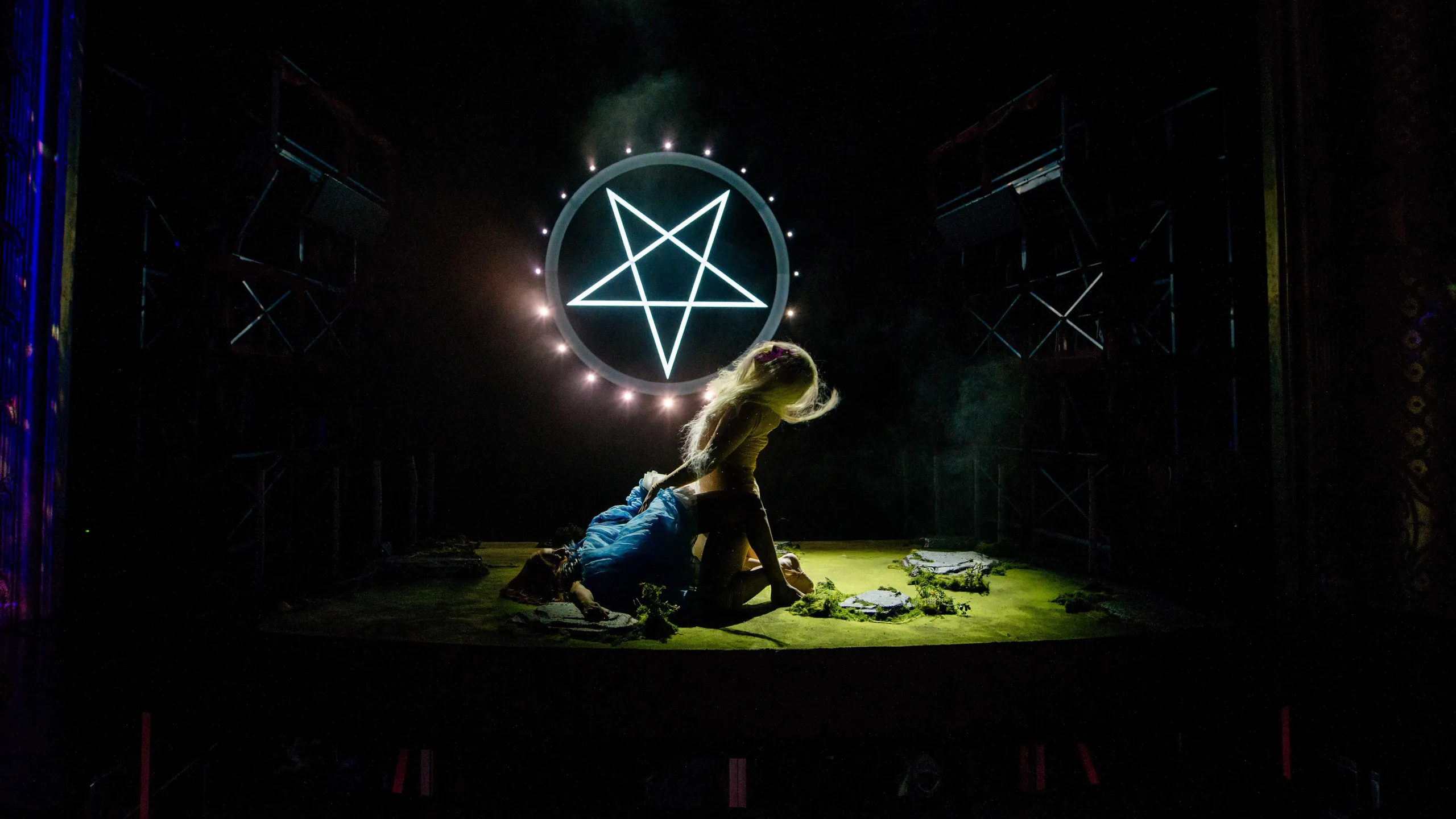 Person med langt lyst hår sitter over en annen i blå kjole foran en lysende pentagramring, i mørk og tåkete teaterscene.